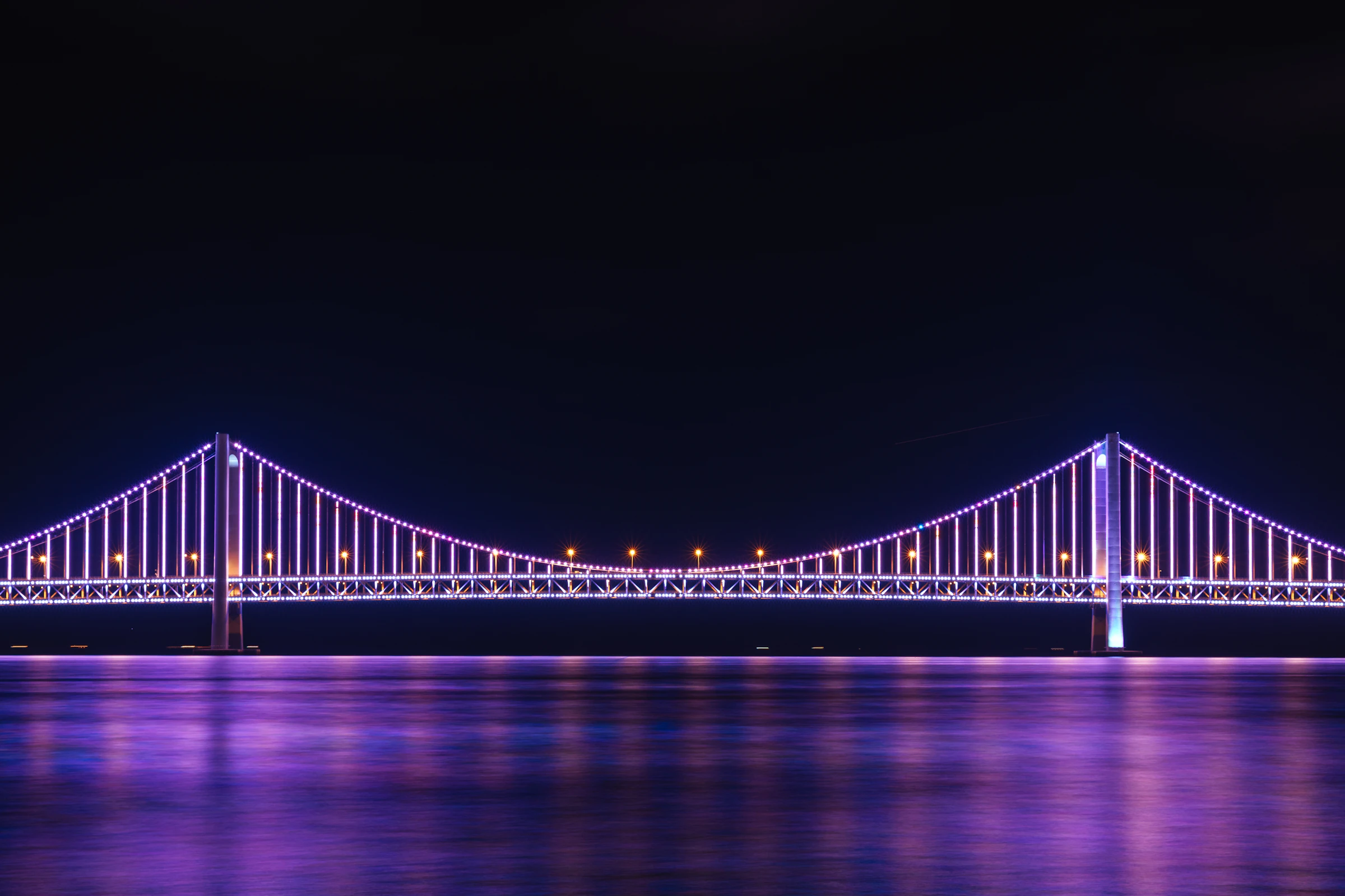 The Gwangan Bridge lit up in purple and blue over the water at night, seen from Gwangalli Beach in Busan