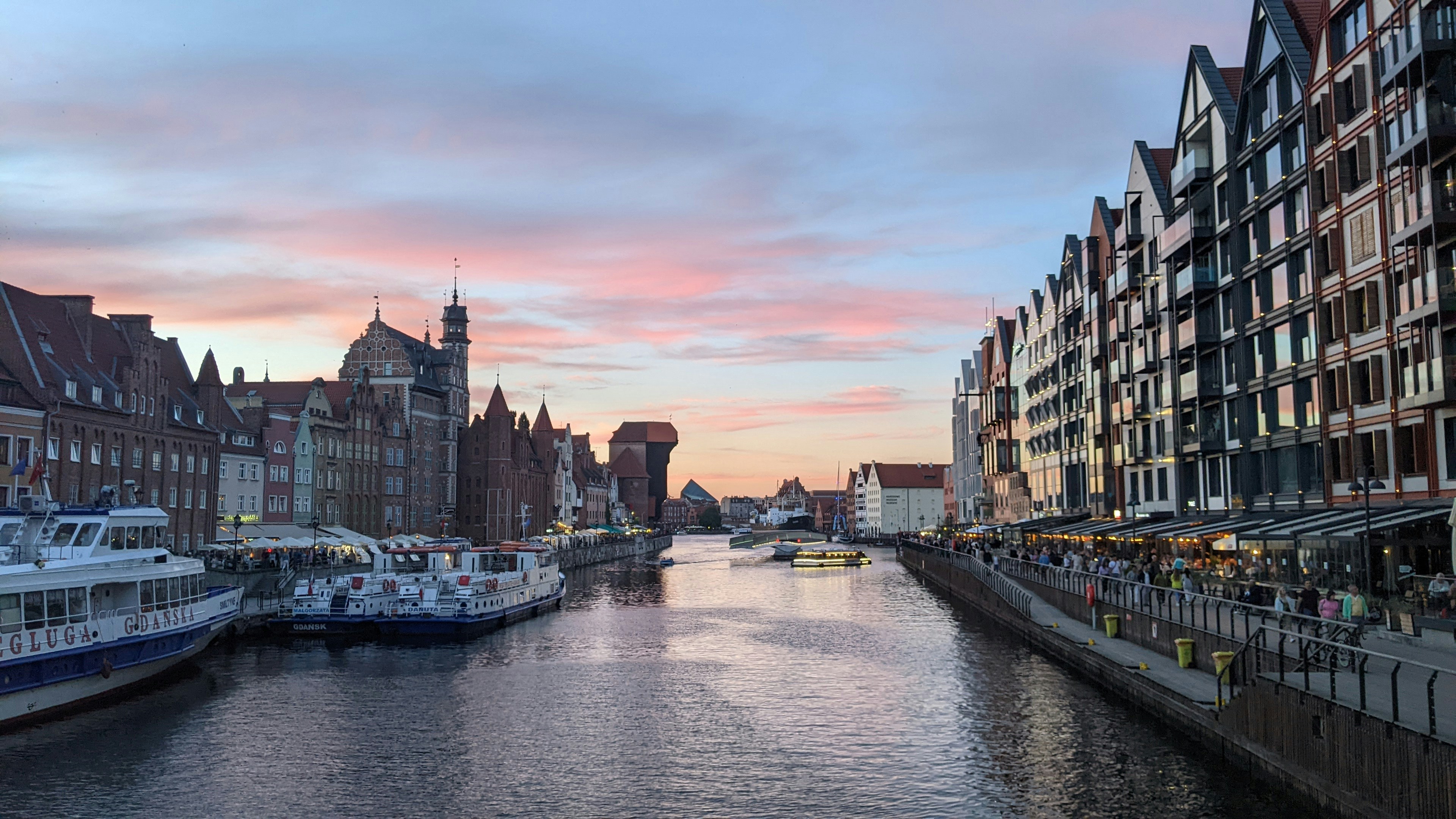 A river running through a city next to tall buildings, A picturesque view of the historic city of Gdansk, Poland. The colorful, medieval buildings line the waterfront, reflecting in the calm waters of the Motława River. A beautiful sunset casts a warm glow over the city, creating a truly magical atmosphere.