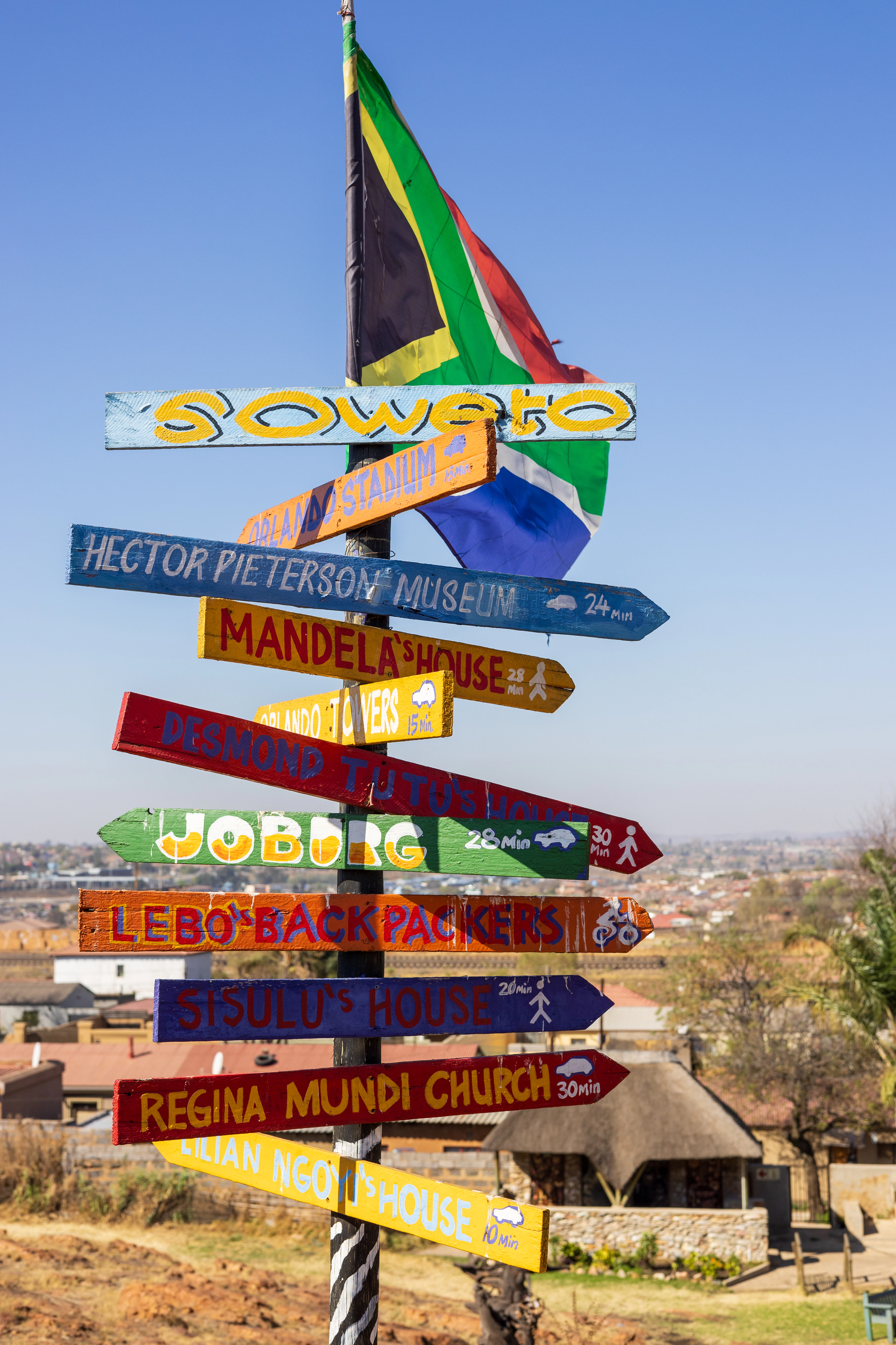 A hand-painted Soweto signpost under the South African flag, its stacked wooden arrows pointing to the Hector Pieterson Museum, Mandela's House, Orlando Towers, Desmond Tutu's house and Regina Mundi Church, with the township's rooftops spread out below in bright winter light.