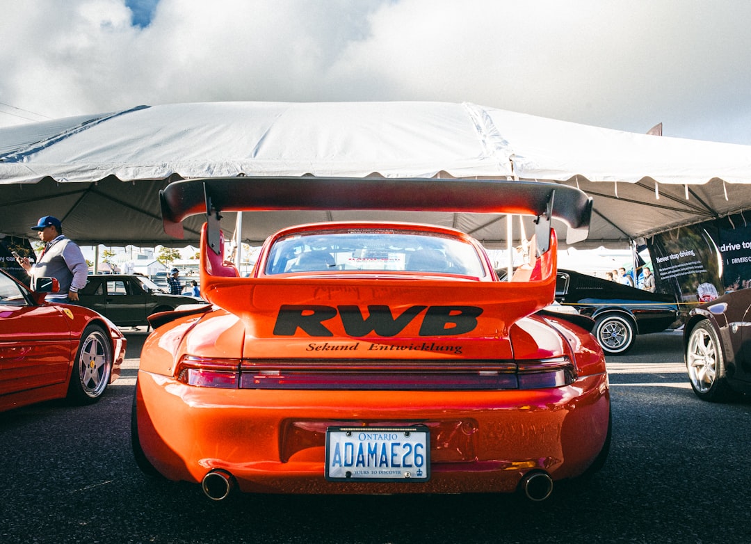 A group of cars parked next to each other in a parking lot, This striking shot captures the rear of a bright red RWB Porsche, with its oversized wing and bold RWB branding. The tent canopy and other vehicles in the background add to the event atmosphere, while the vivid color and stance of the Porsche command attention, celebrating the tuning culture and unique styling of RWB.