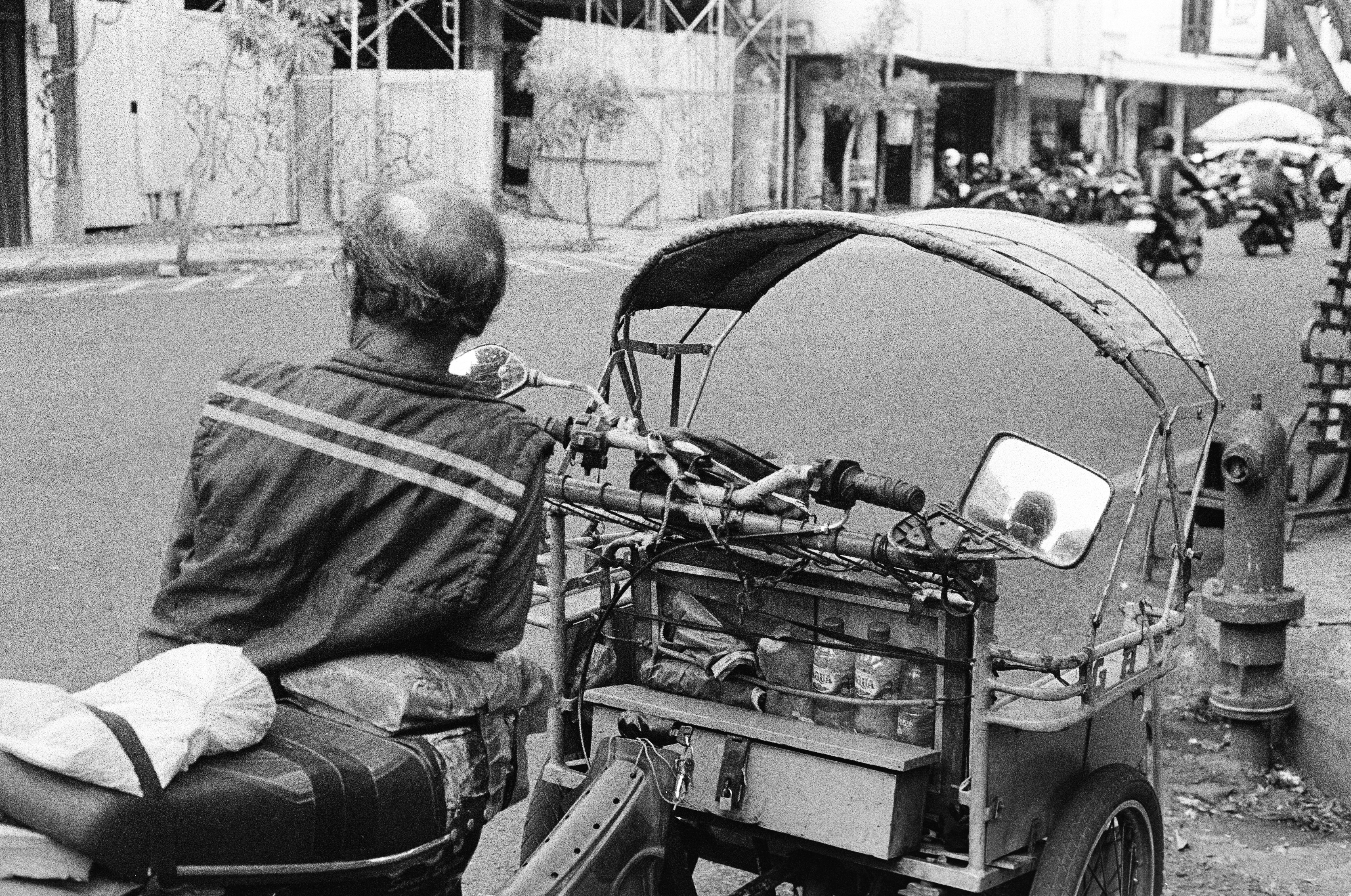 A man riding on the back of a motorcycle down a street, An oldman sits leaning on his bentor (modified pedicab), taken with Nikon FM2n using Ilford HP5+ 400 film.