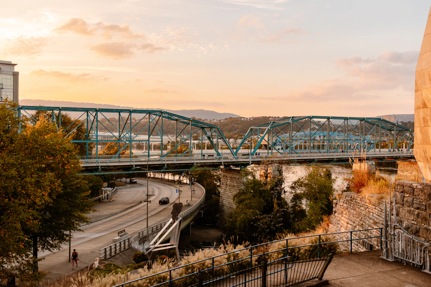 A truss bridge crossing a broad river at sunset, with the city along the bank.