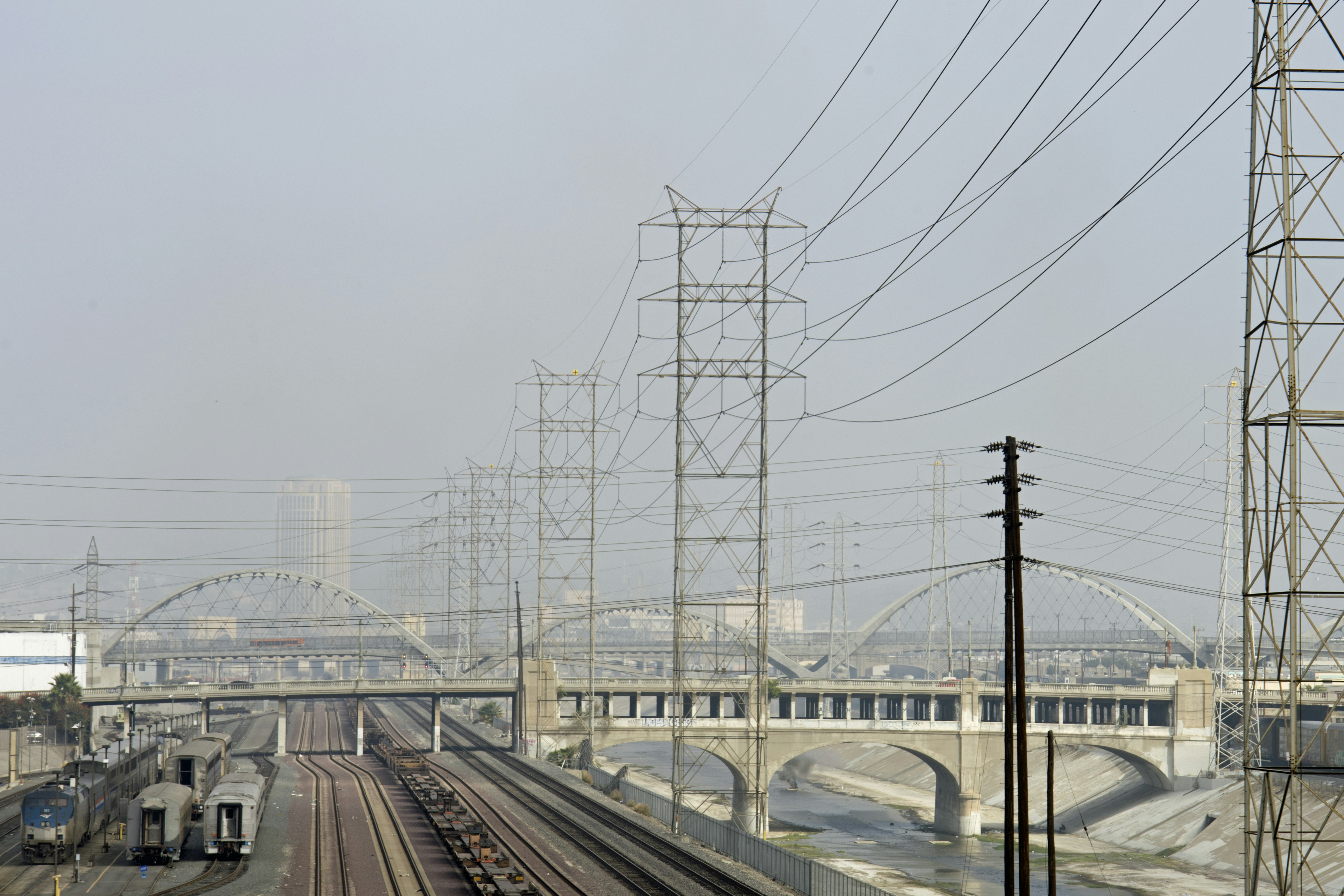 A train traveling down train tracks next to a bridge, A gritty view of the Los Angeles River and surrounding train tracks, bridges, and electricity towers and wires.