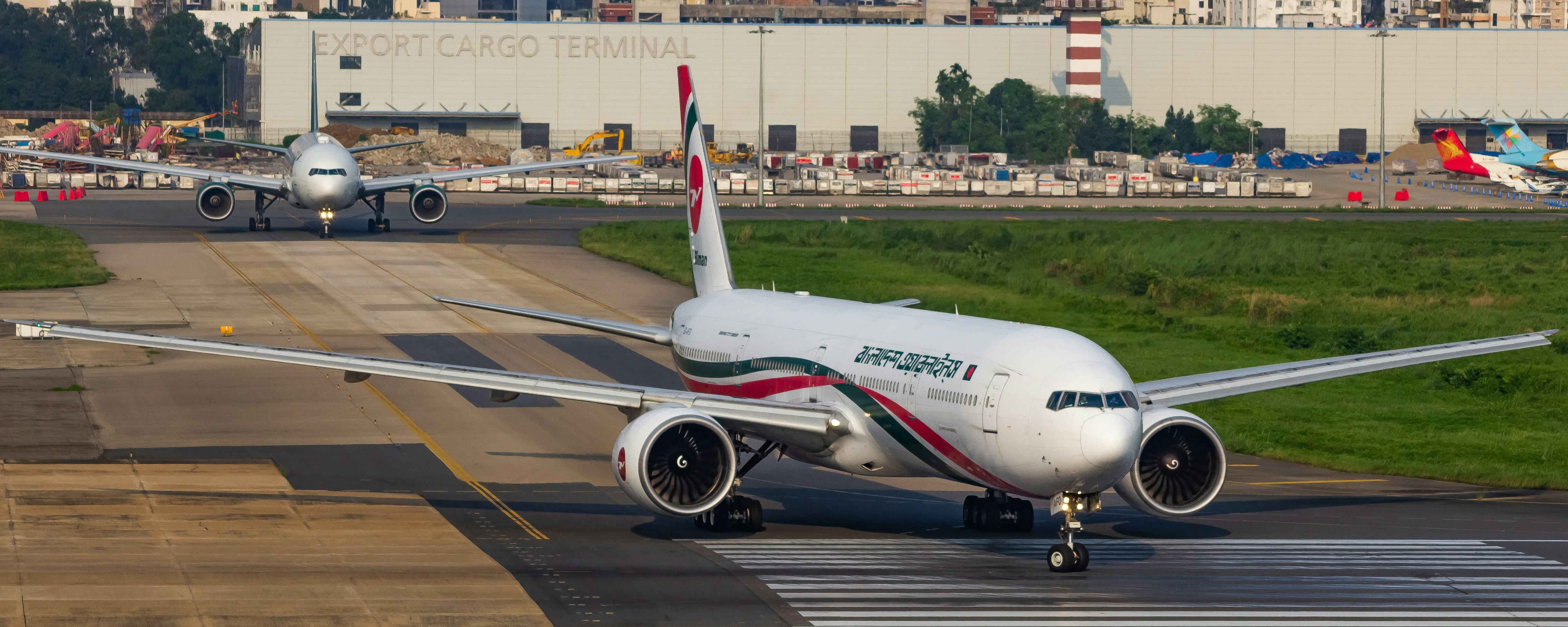 Two airplanes taxi on an airport tarmac near cargo terminal buildings.