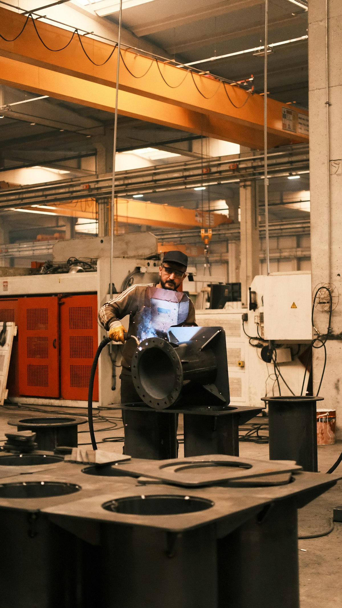 A welder in protective gear works on a large black steel fabrication in a bright shop with an orange overhead crane spanning the ceiling.