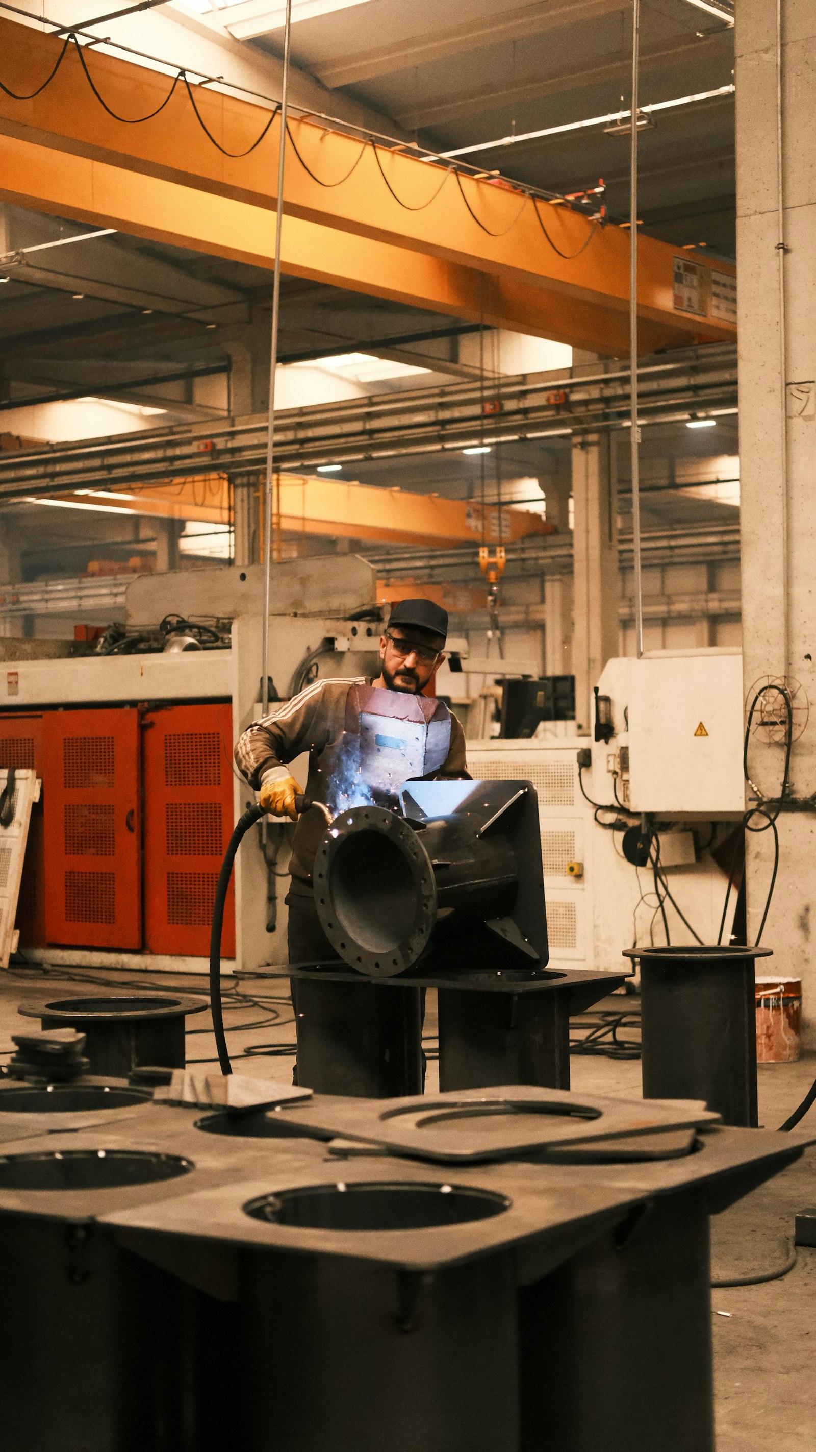 A welder in protective gear works on a large black steel fabrication in a bright shop with an orange overhead crane spanning the ceiling.