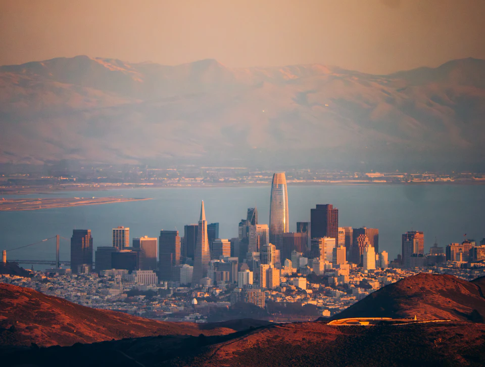 The image presents a panoramic view of the San Francisco Bay Area, with the city's skyline visible from a high vantage point. The bay is nestled between mountains and hills, adding depth to the scene. The cityscape features numerous buildings in various sizes and colors, including a tall skyscraper that stands out among them. The sky above is painted in shades of orange and pink, suggesting either sunrise or sunset, casting a warm glow over the entire scene.

The image does not contain any discernible text or countable objects, making it an open-air view of the city's skyline.