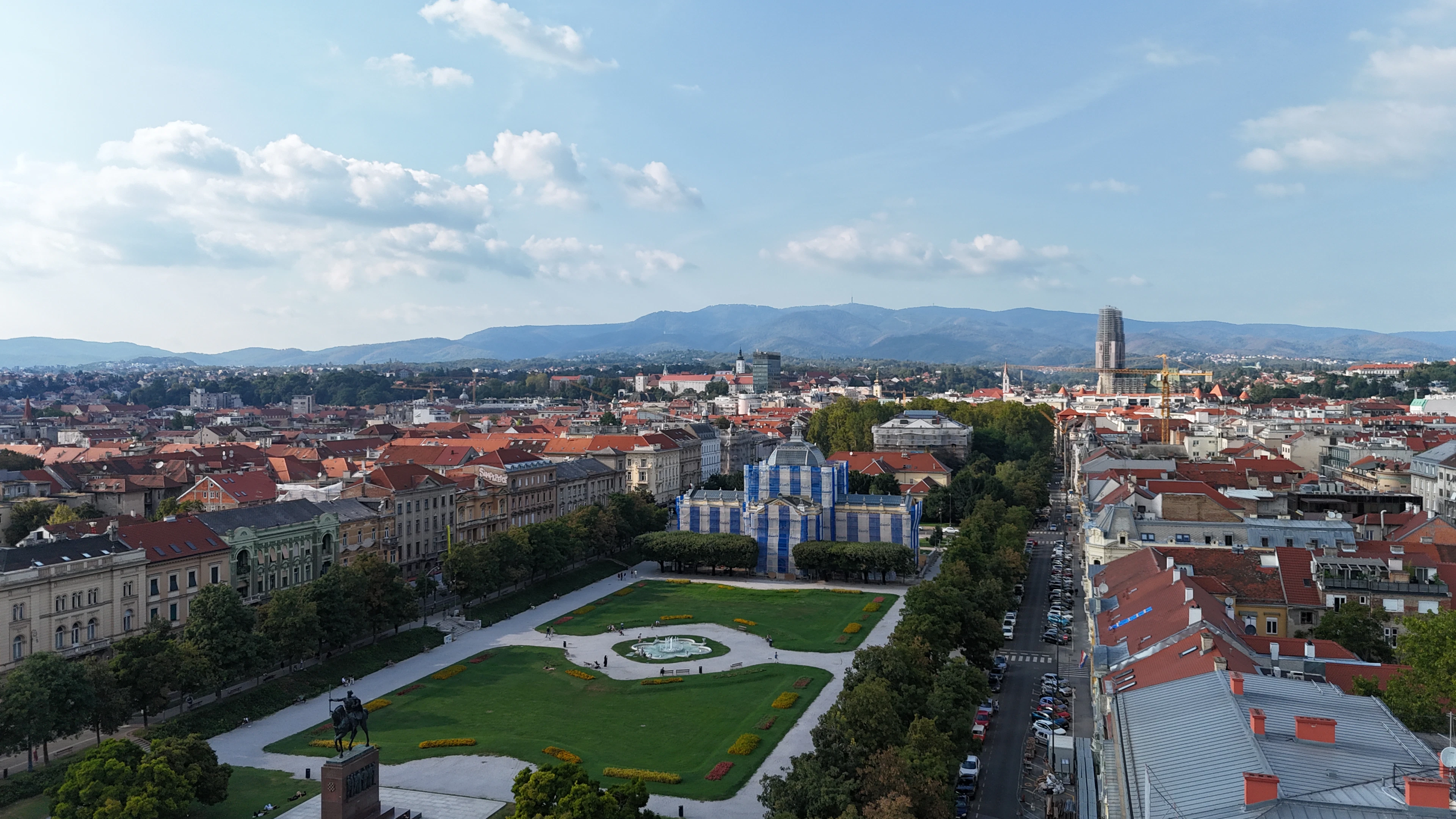 Aerial view of a formal park with a fountain and pavilion, flanked by red-roofed buildings and a distant slender spire.