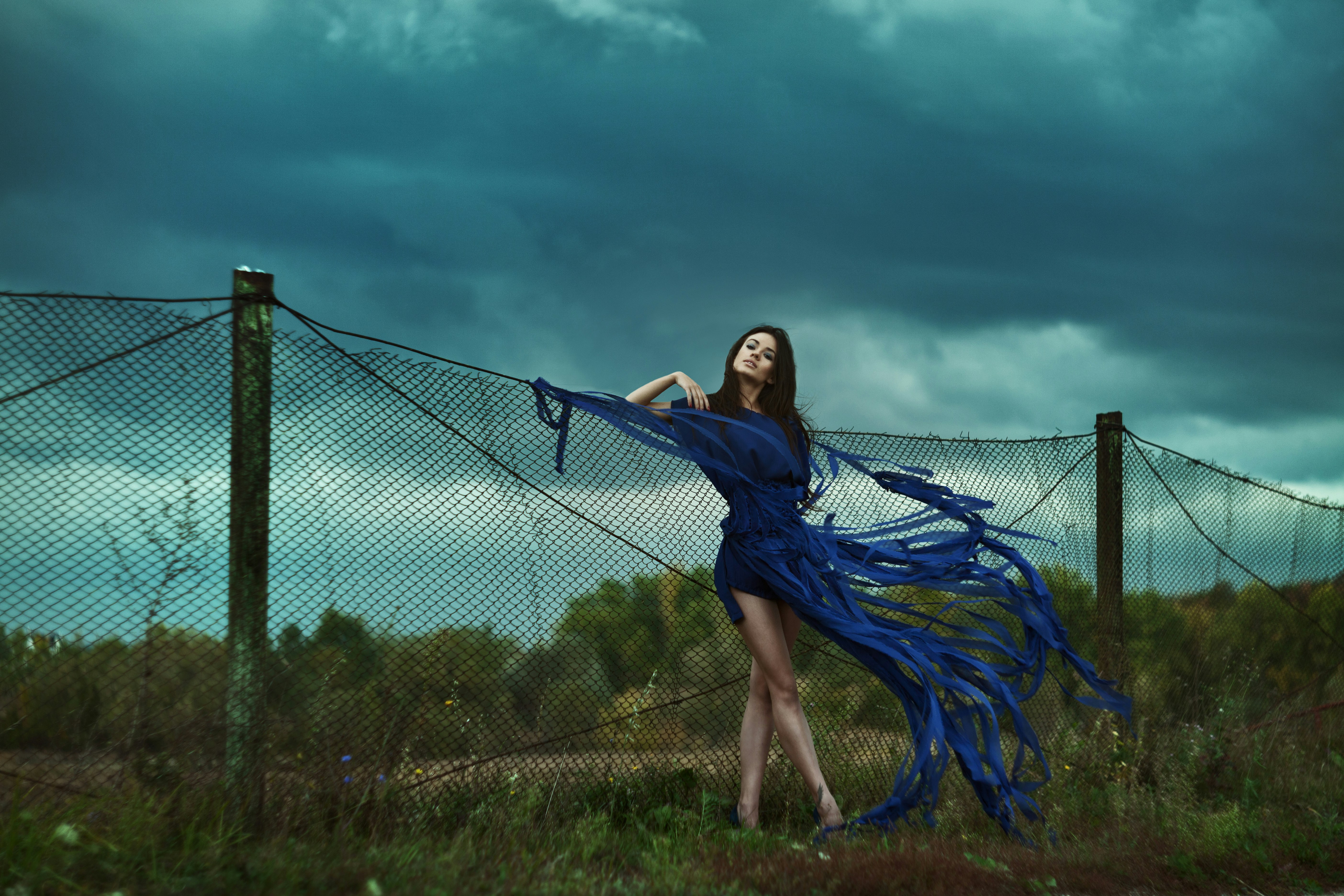 Une femme contre un grillage laisse voler sa robe bleue dans le vent.