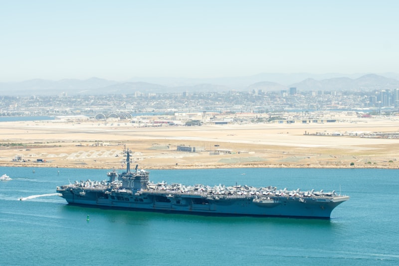 A US Navy aircraft carrier sailing in open water with a city skyline in the background