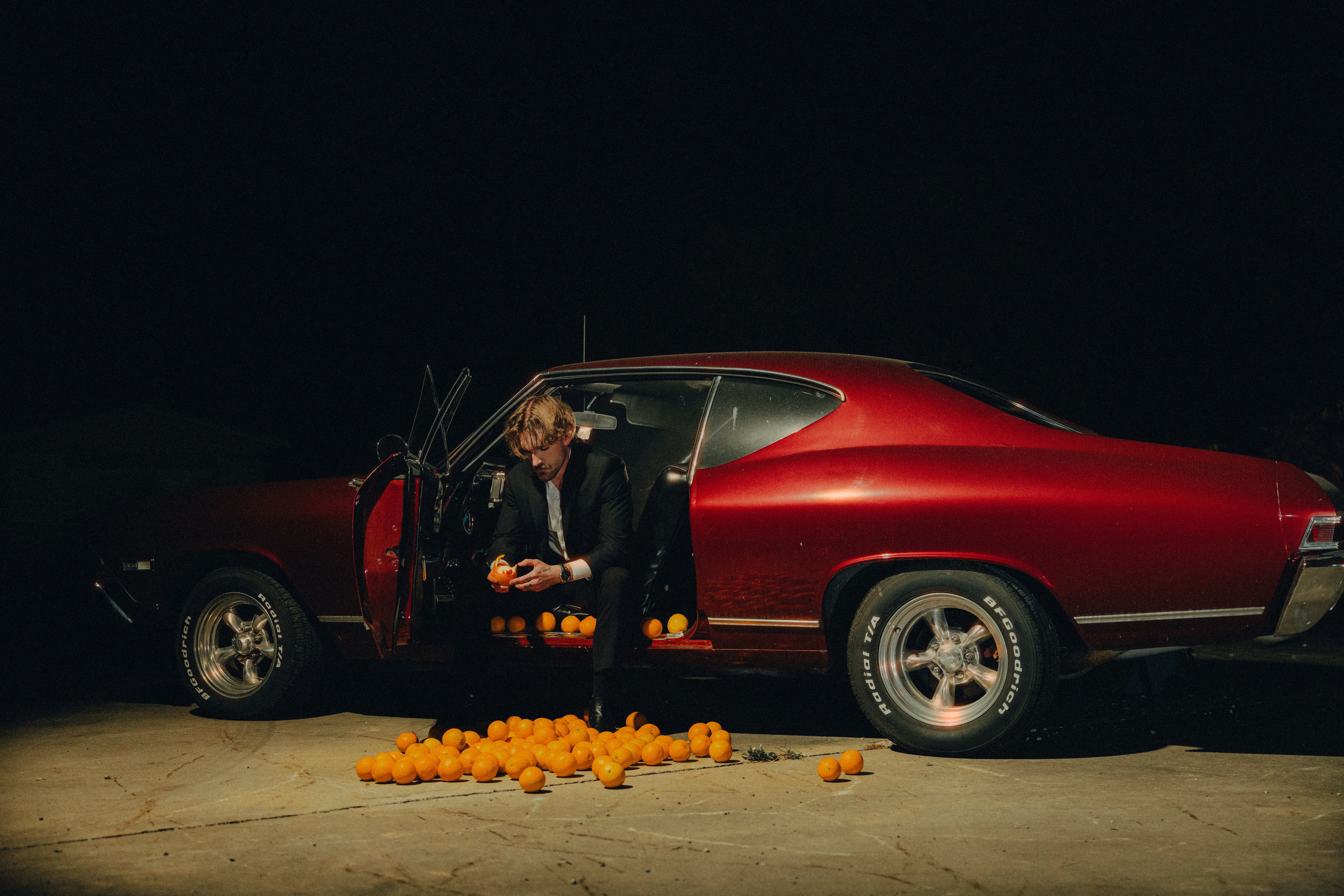A man standing next to a red car filled with oranges
