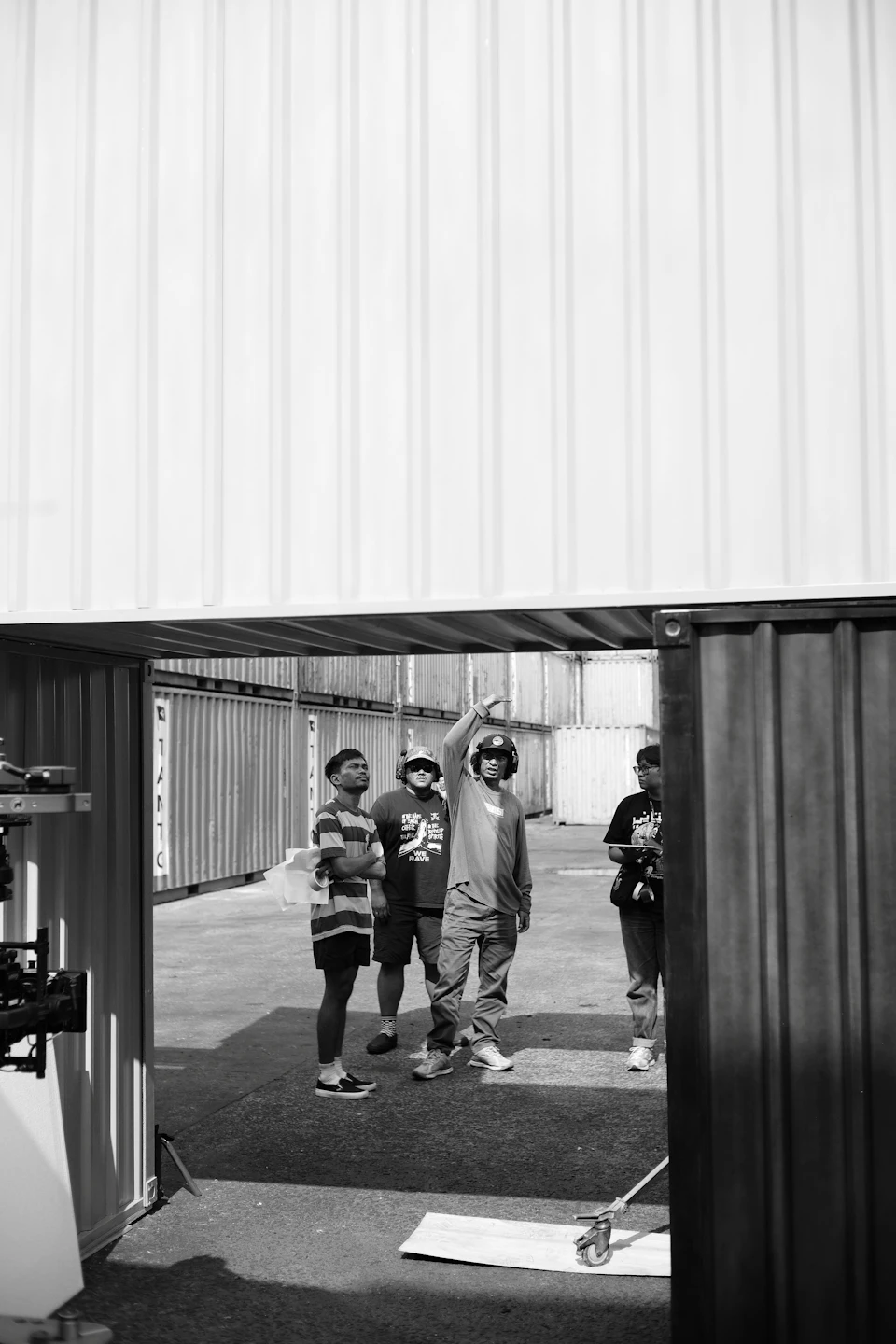 Four people stand on concrete, discussing or observing outside a large white shipping container