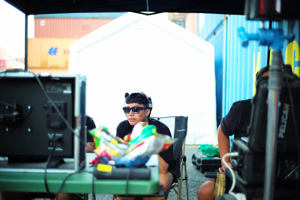 The image shows a person sitting on a green chair, wearing sunglasses and holding a bag with various items inside. The person is seated under an umbrella-like structure that appears to be white or gray in color. In the background, there are two other people present - one standing behind the person sitting on the chair and another person sitting further back. A television screen can also be seen in the scene, suggesting a possible filming location or outdoor event.