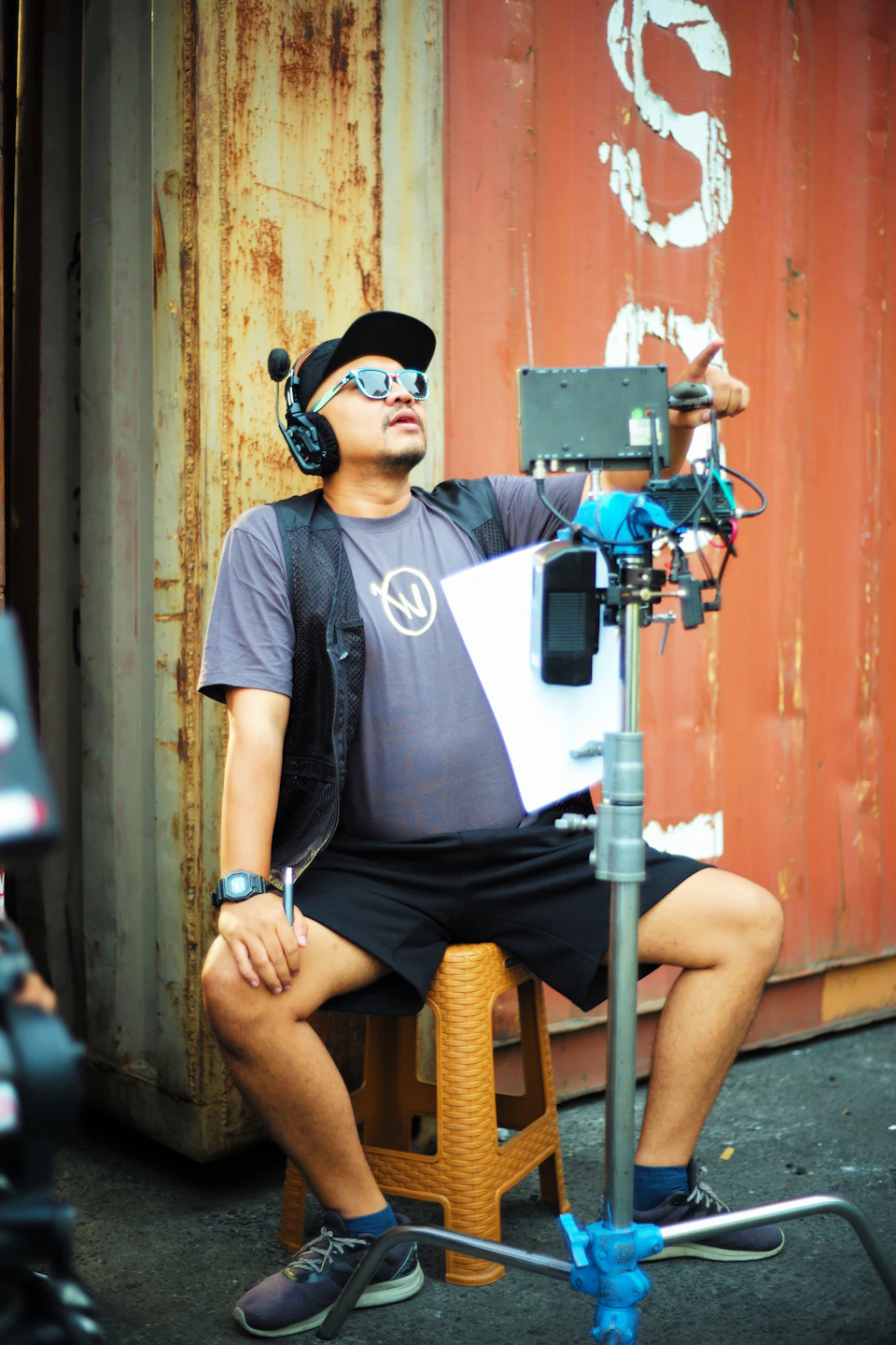 Man seated on orange stool, wearing gray t-shirt and black shorts, holding camera