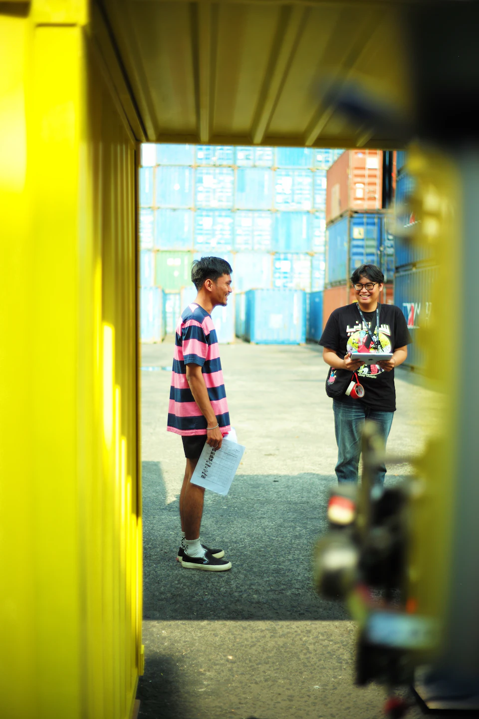 The image shows two young men standing on a concrete floor, engaged in conversation. One man is holding a piece of paper and appears to be reading it while talking to his companion. The other man is holding a camera, possibly taking photographs or recording the interaction between them. In the background, there are several shipping containers stacked up against a yellow wall, suggesting an industrial setting.