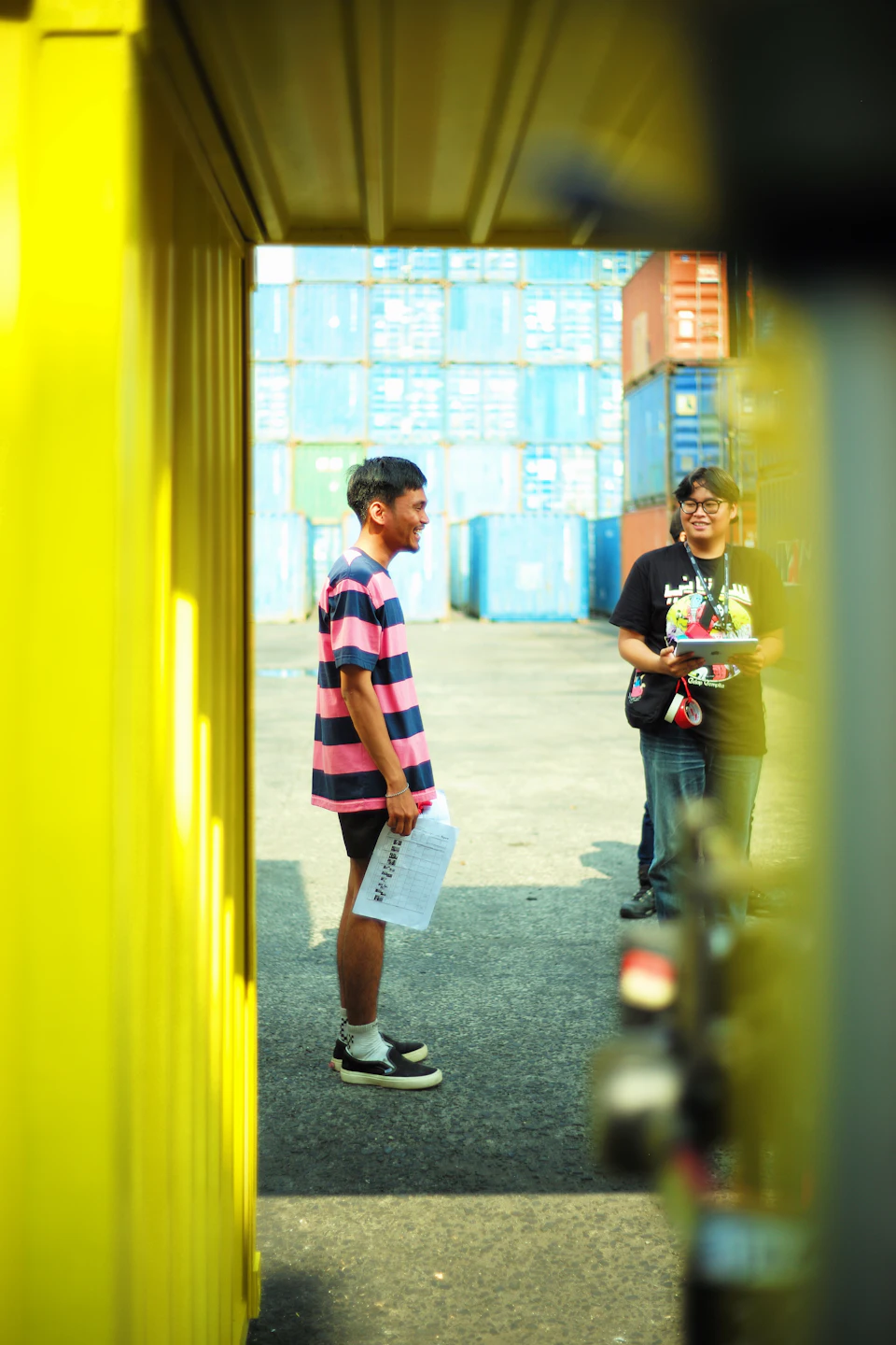 The image shows a man and a woman standing on a sidewalk, engaged in conversation. The man is wearing a striped shirt with pink and blue stripes, while the woman has black hair and is dressed in a black t-shirt. They are positioned next to a yellow building that appears to be a shipping container or warehouse. A motorcycle can also be seen parked nearby.
