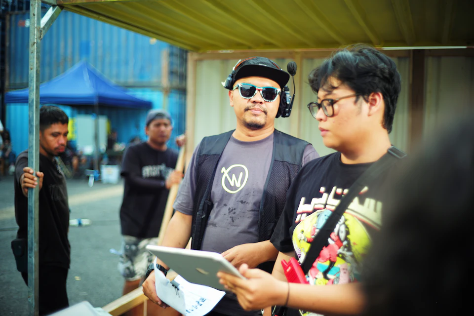 The image shows a group of people gathered around an outdoor event, with two men prominently featured. One man is holding a tablet computer and appears to be explaining something to his companion who is wearing a black t-shirt adorned with a graphic design. The other man in the scene is also wearing a black t-shirt but has a different design on it. In the background, there are several people standing under a yellow canopy or tent, suggesting that this event might be taking place outdoors during an outdoor festival or gathering.