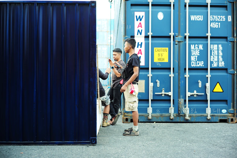 The image shows three people standing near a blue shipping container with white lettering on it, which reads "WASSU 57525 U". The person on the left is holding a cell phone and appears to be engaged in a conversation or taking a photo. In the center of the frame, another individual is wearing a black shirt and khaki pants, while the third person stands behind them with their back turned towards the camera.

In the background, there are two more shipping containers visible - one on the left side and another on the right side of the image.
