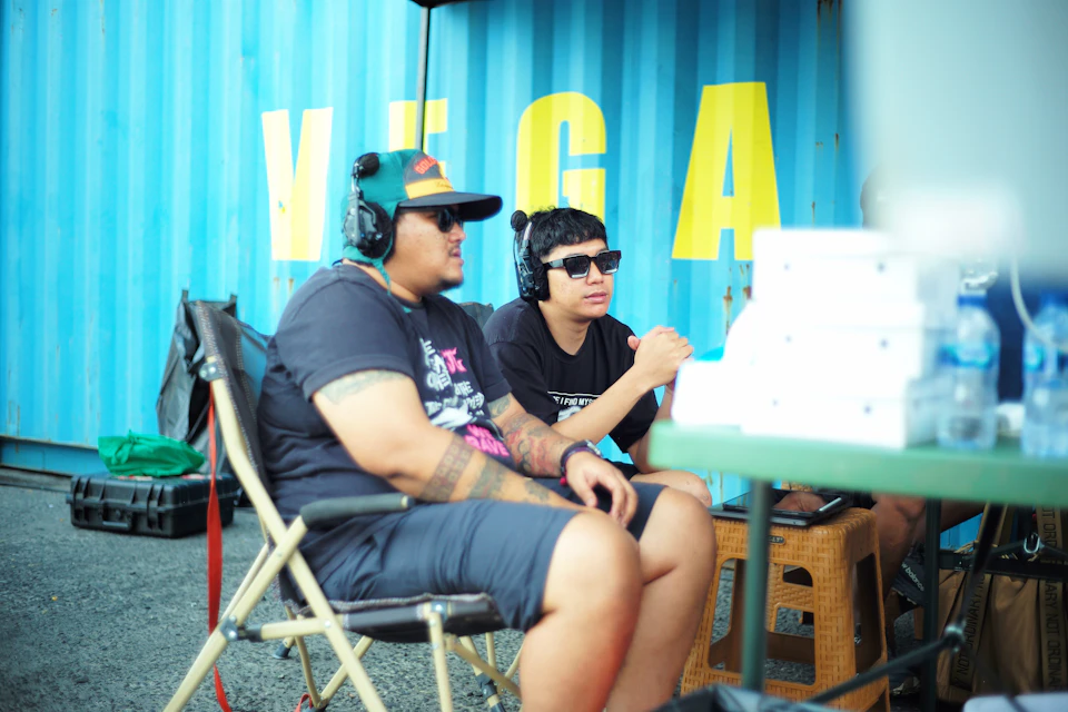 The image shows two men sitting on a green folding chair, both wearing headphones and sunglasses. They are seated at a table with a white box placed on it. The background features a blue wall with the word "VEGA" written in yellow letters.