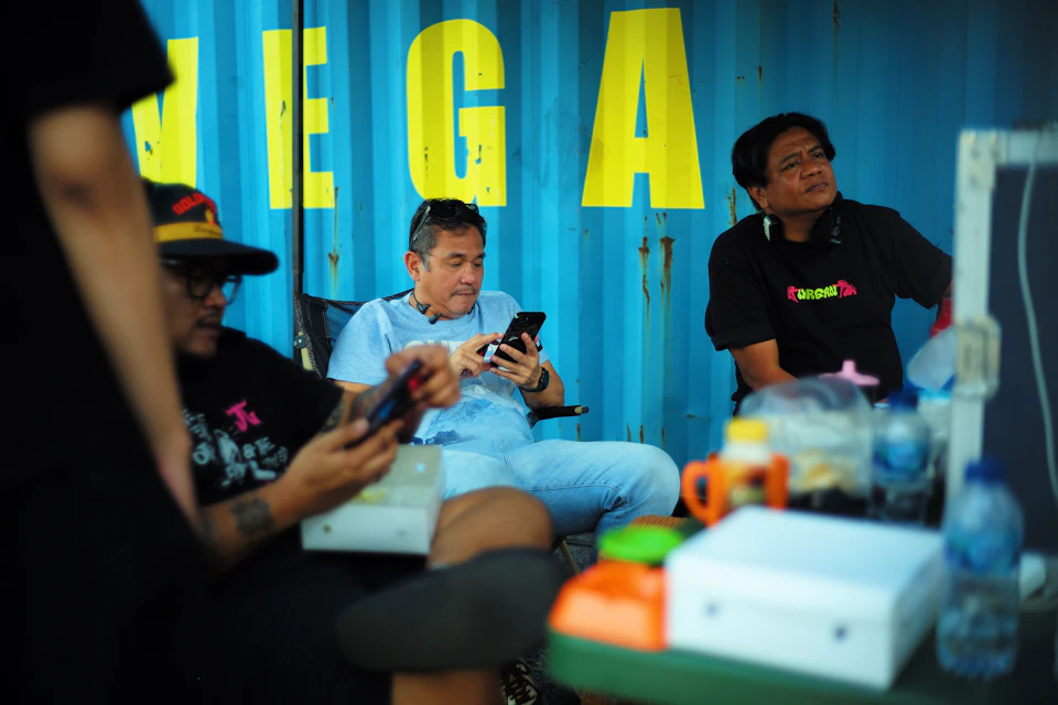 The image depicts a group of people gathered around a table, engaged in various activities. The main focus is on three individuals seated at the table, with one person using their phone and another two looking up towards the sky. The background features a blue wall adorned with yellow text that reads " mega". On the table, there are several items including water bottles, cups, and a sandwich.