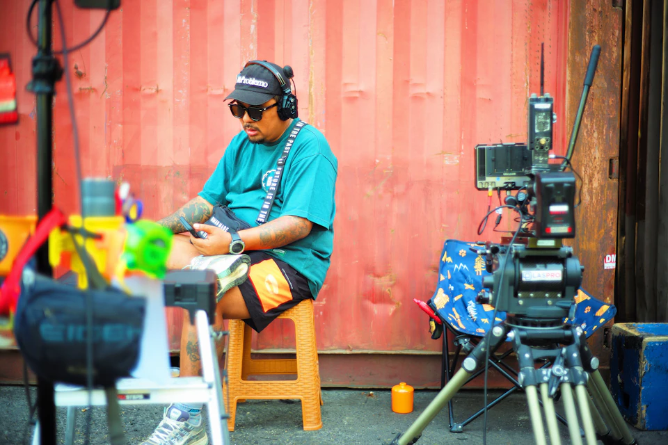 In the center of a bustling city, a man is seated on an orange stool with his legs crossed and wearing a blue t-shirt and black sunglasses. He holds a phone to his ear, suggesting he might be engaged in a conversation or listening to music. Behind him, there are two large cameras mounted on tripods, indicating that this scene could be related to photography or filming. The backdrop is a red wall with a window, adding an urban touch to the setting.