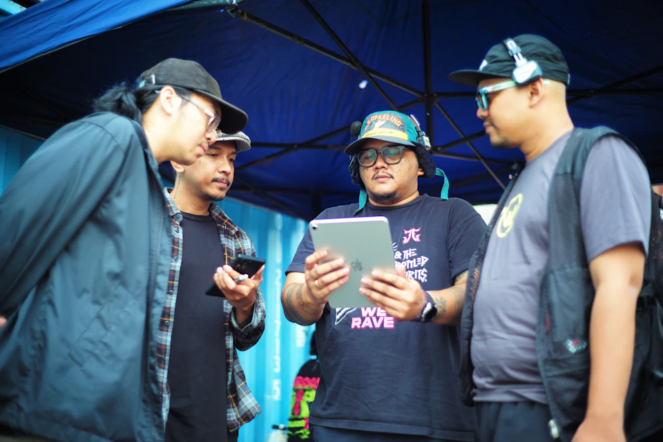 In the image, there are four men standing under a blue tent on what appears to be an outdoor event or gathering. The man at the center is holding up a tablet computer and showing it off to his friends. He's wearing a black baseball cap with a colorful design. To his left, another man is also holding a tablet device, while two more men stand nearby, one of whom is wearing a black t-shirt.

The scene takes place in an outdoor setting, possibly at a festival or similar event, as suggested by the presence of the tent and the casual attire of the people present.