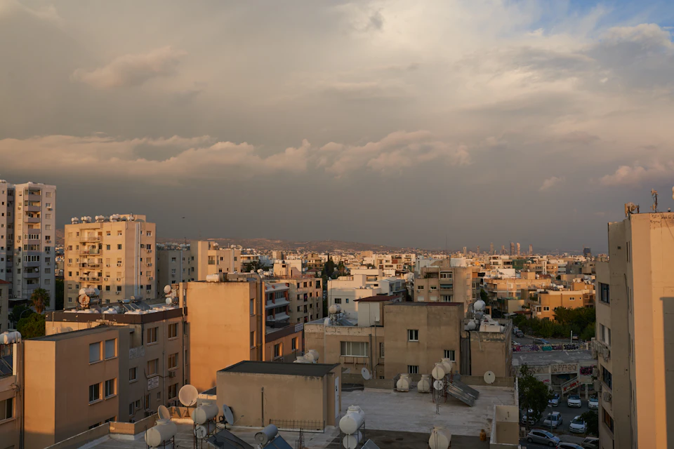 The image presents a cityscape viewed from an elevated perspective, with buildings of varying heights and designs stretching out into the distance under a cloudy sky. The tallest building is situated on the left side of the frame, while the shortest one can be seen on the right. The buildings are predominantly white or light-colored in color, creating a sense of uniformity across the cityscape. A few cars are parked along the street below, adding a touch of everyday life to this urban scene.