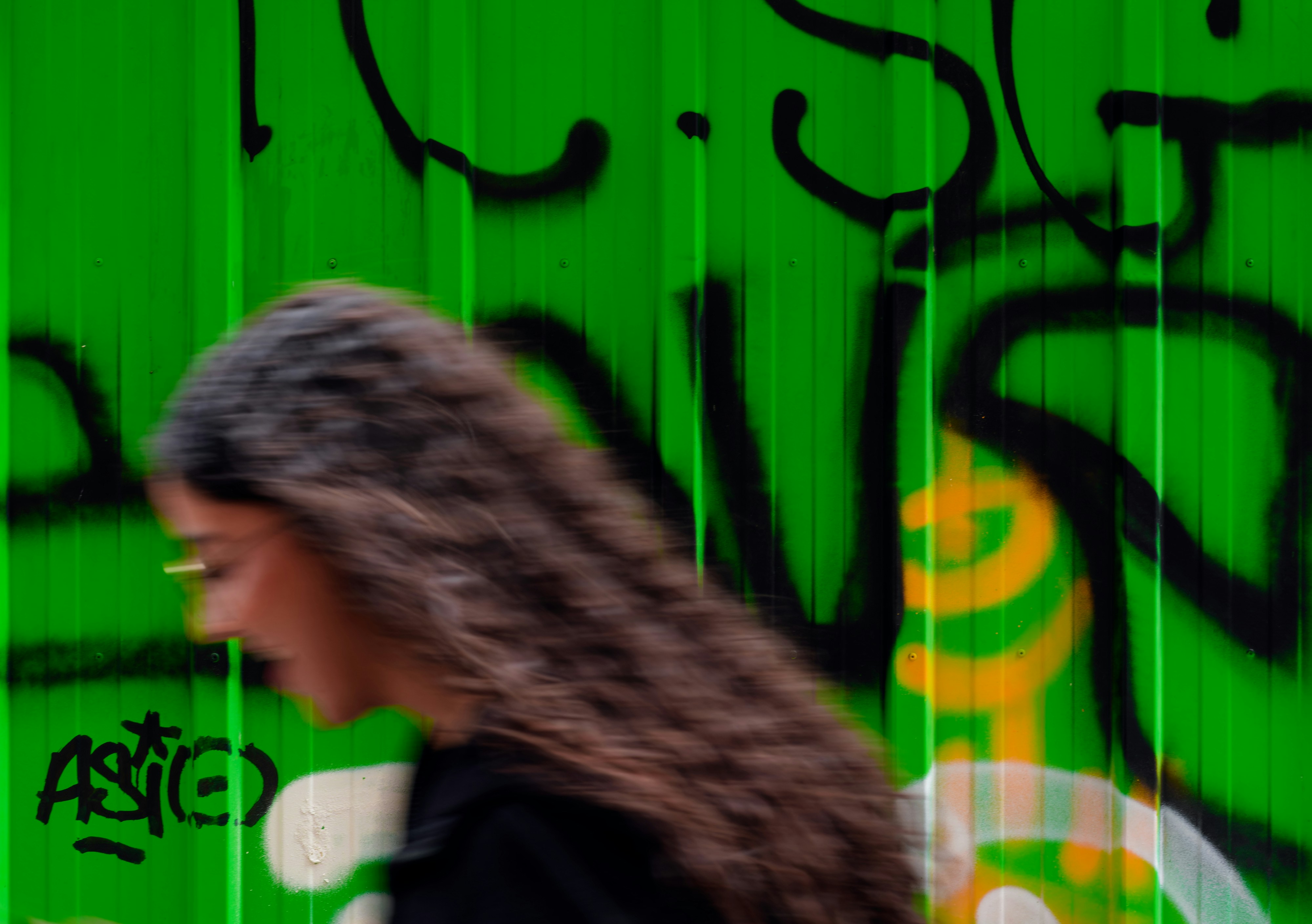 A woman walking past a green wall covered in graffiti