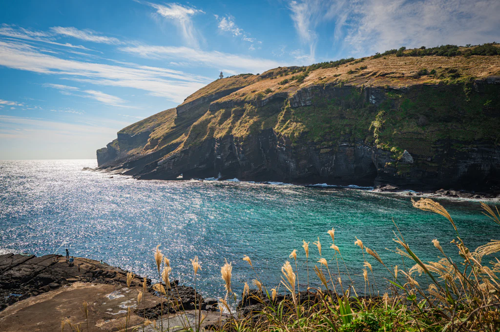 Turquoise water below the dark volcanic cliffs of a grass-topped headland on Udo, off Jeju