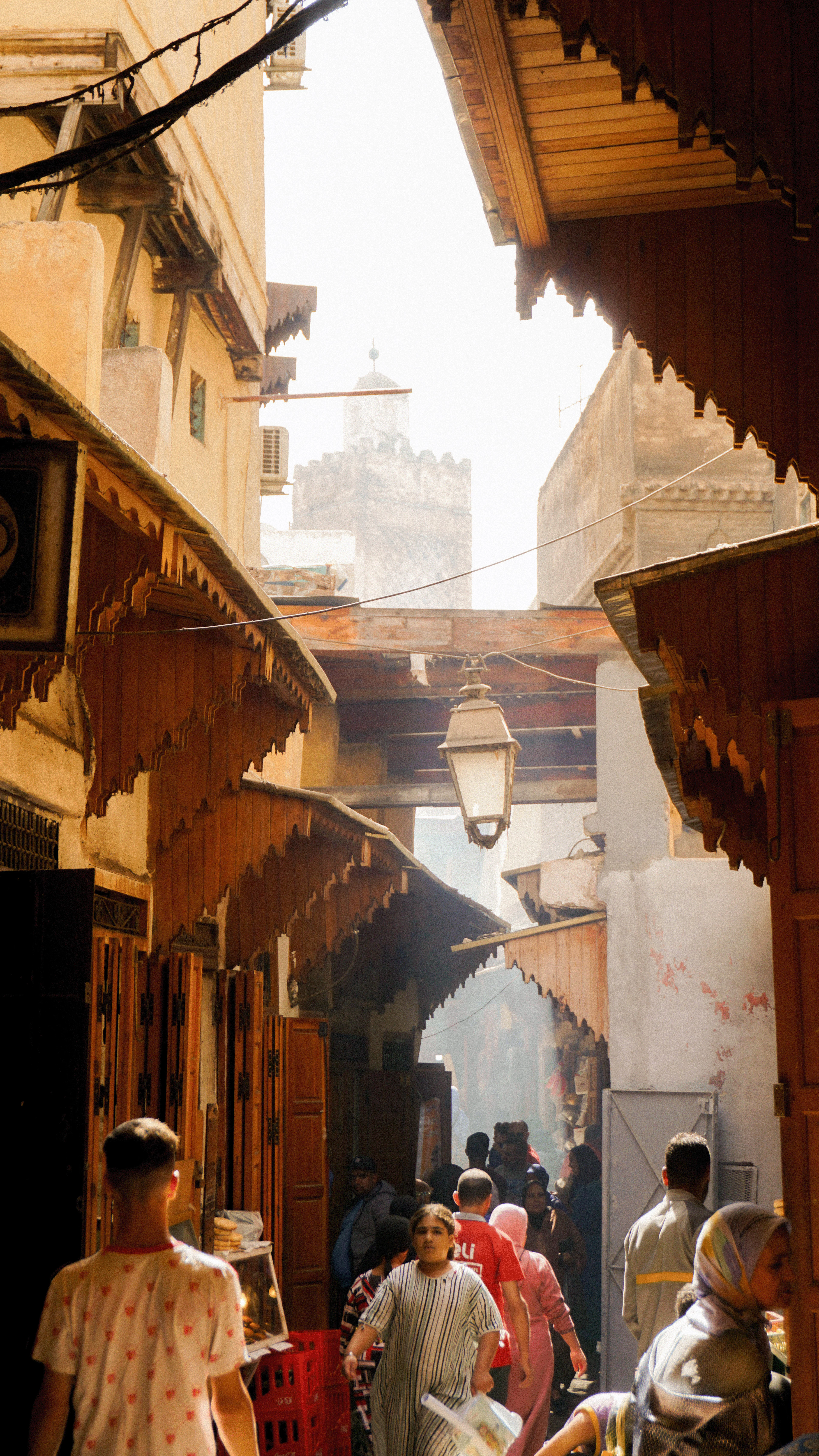 A crowded covered lane in the Fez medina, shoppers passing beneath carved wooden shop eaves and a hanging lantern, with a minaret rising in the haze beyond