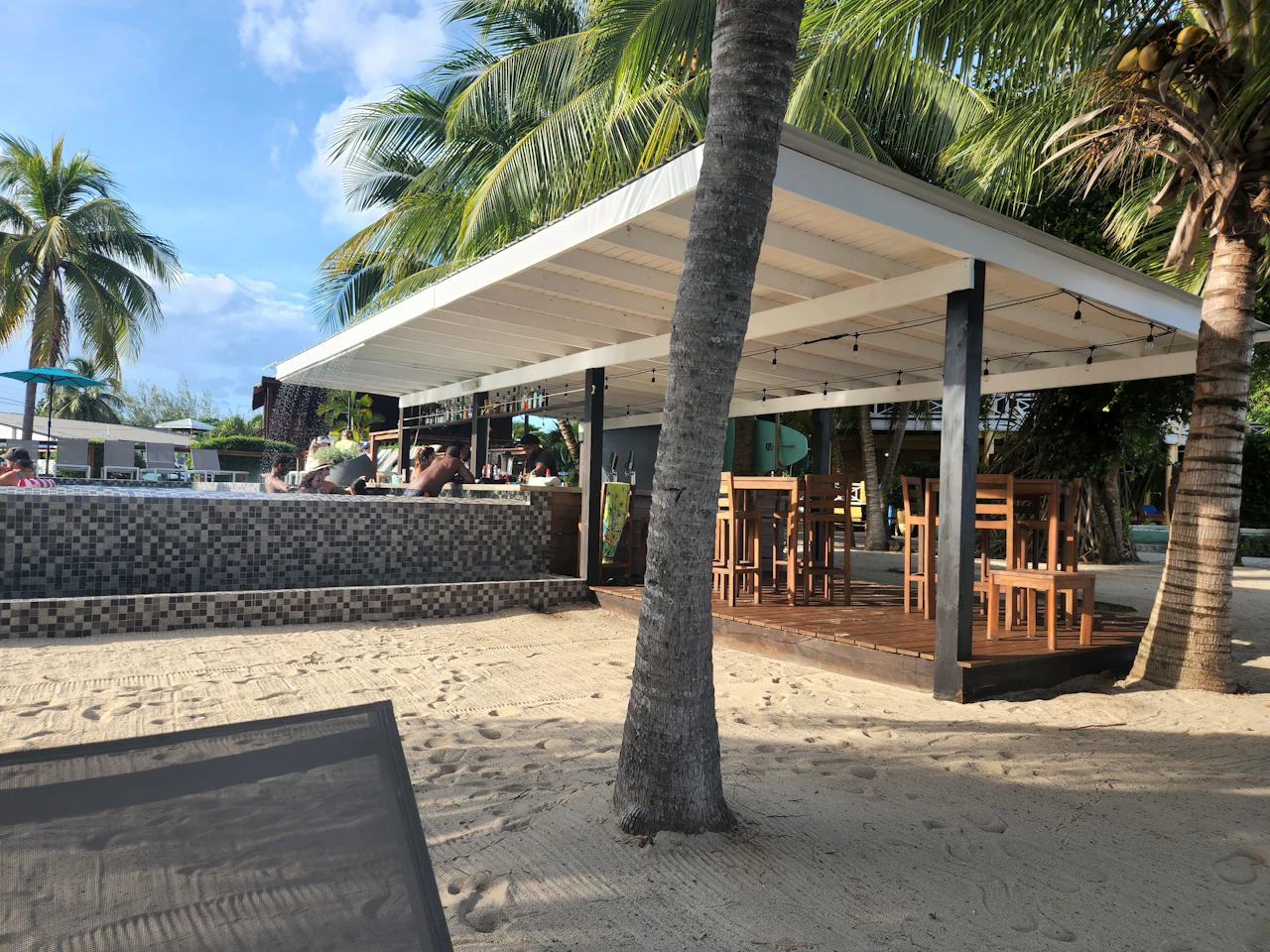 An open-sided beach bar with a white roof among coconut palms, tables and stools on the sand, and a lounger in the foreground.