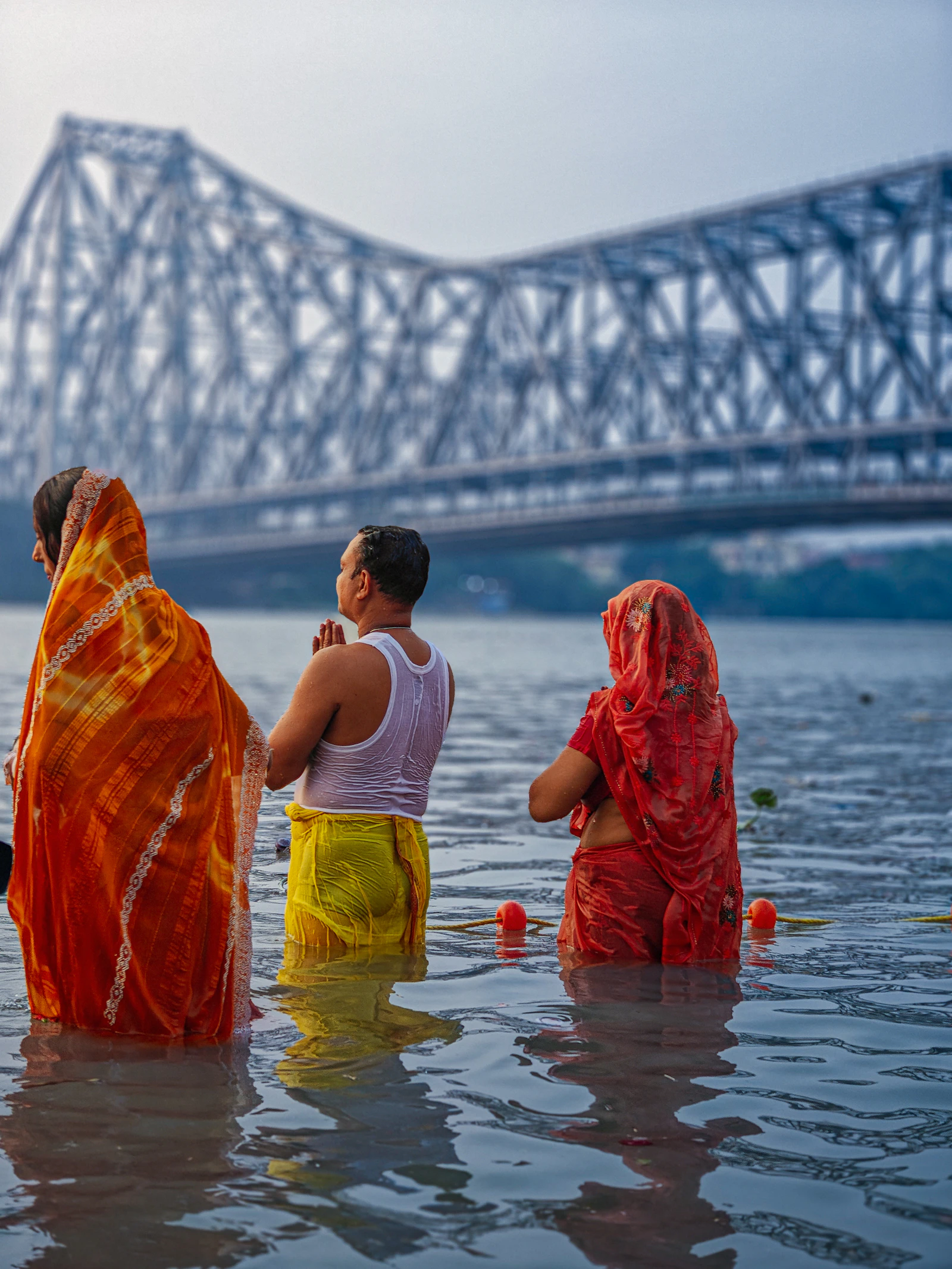 Devotees in colorful clothes stand waist-deep in the Hooghly River in Kolkata with palms joined, the Howrah Bridge behind them