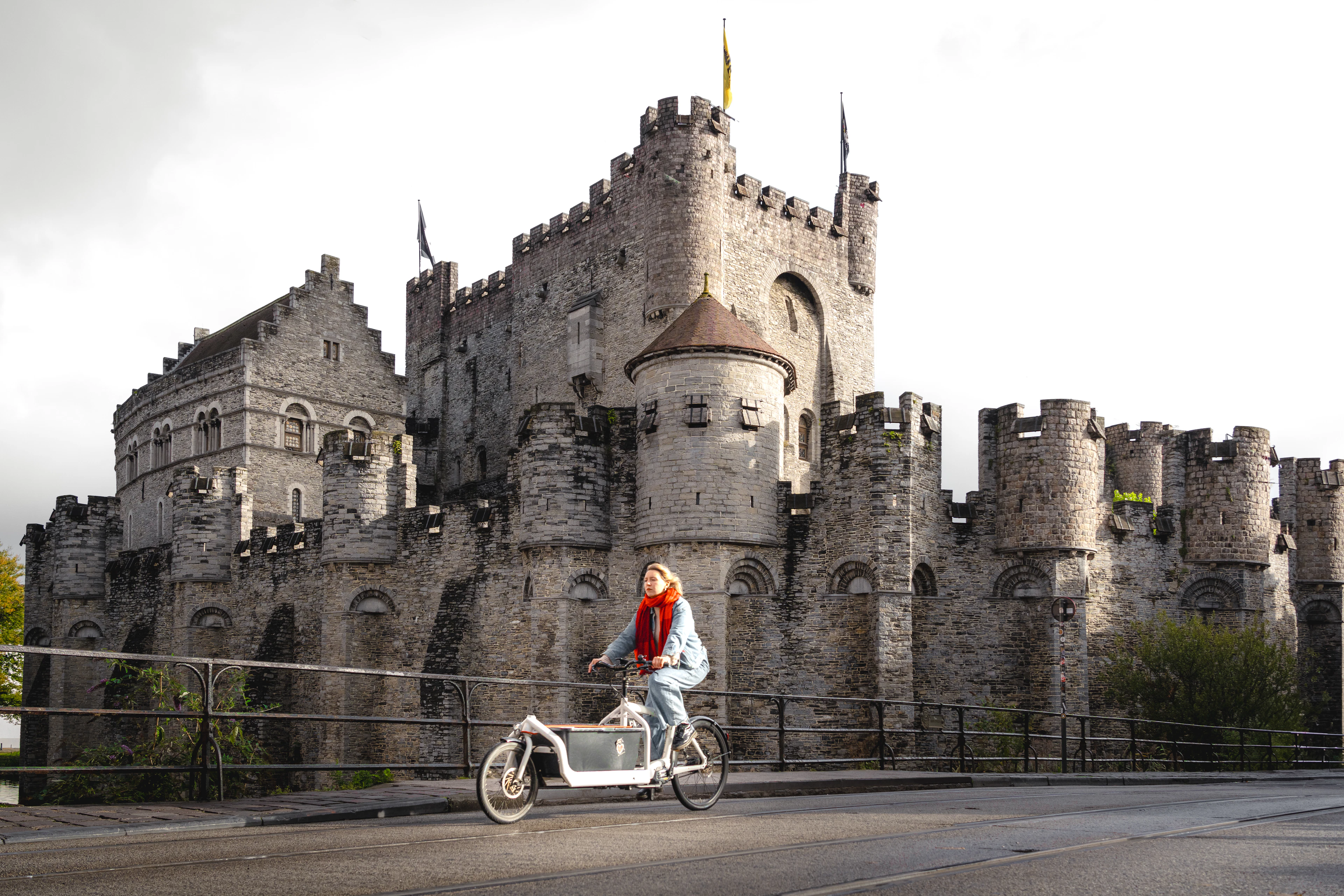 A cyclist in a red scarf crosses the bridge in front of Ghent's turreted Gravensteen castle in soft autumn light.