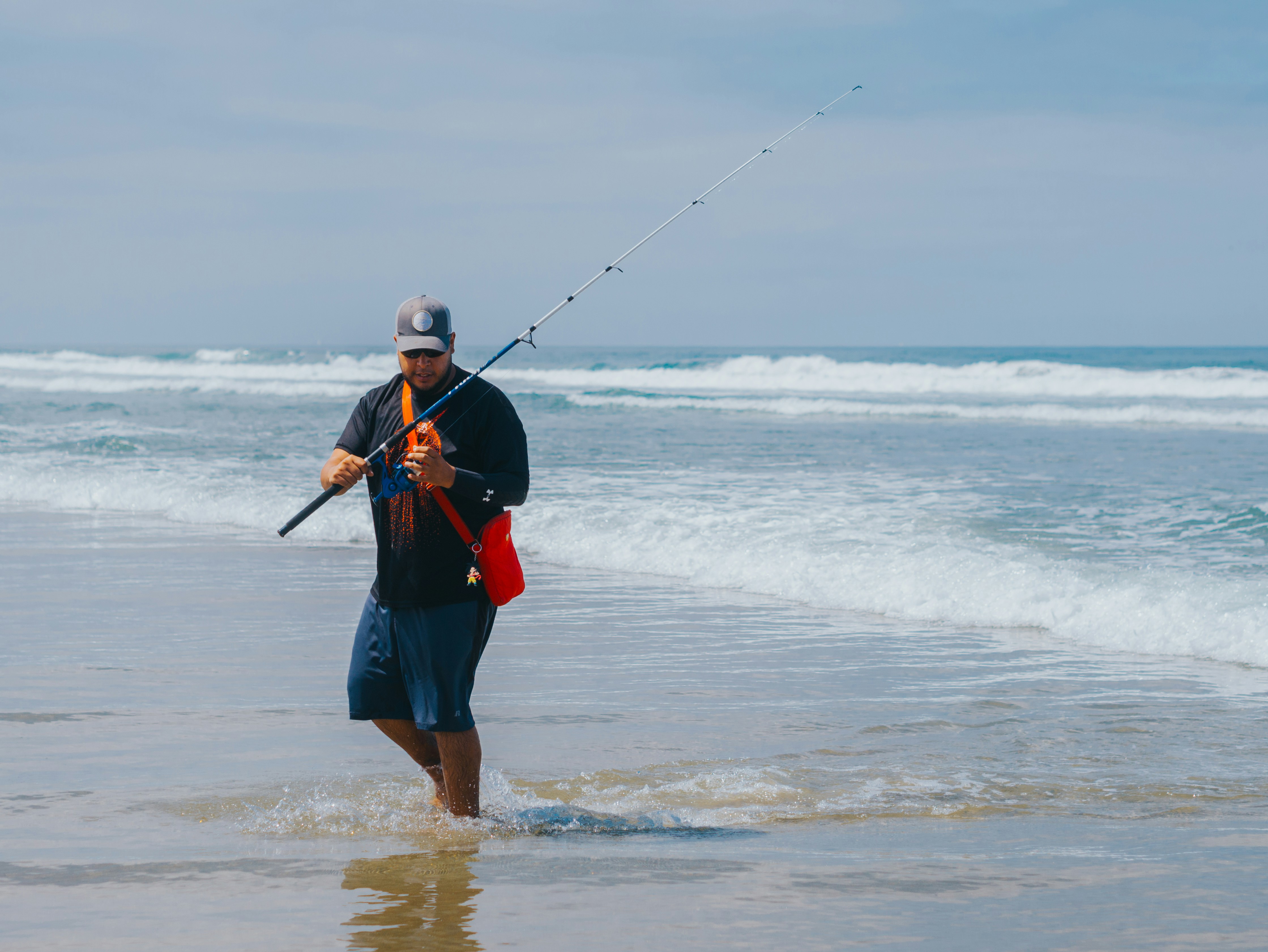 Angler in a black long-sleeve shirt and cap wading out of knee-deep surf with a spinning rod, a red crossbody bag hanging at his hip on an orange strap