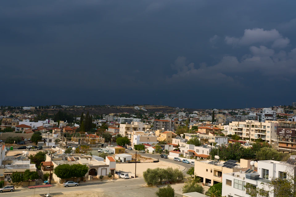 The image presents a panoramic view of a cityscape, captured from an elevated perspective. The sky is filled with dark clouds, suggesting an overcast day or even impending rain. The buildings, varying in height and architectural style, are nestled closely together, creating a dense urban environment. The street below is lined with trees, providing some greenery amidst the concrete structures. Cars can be seen parked along the streets, indicating that this city is active and bustling.