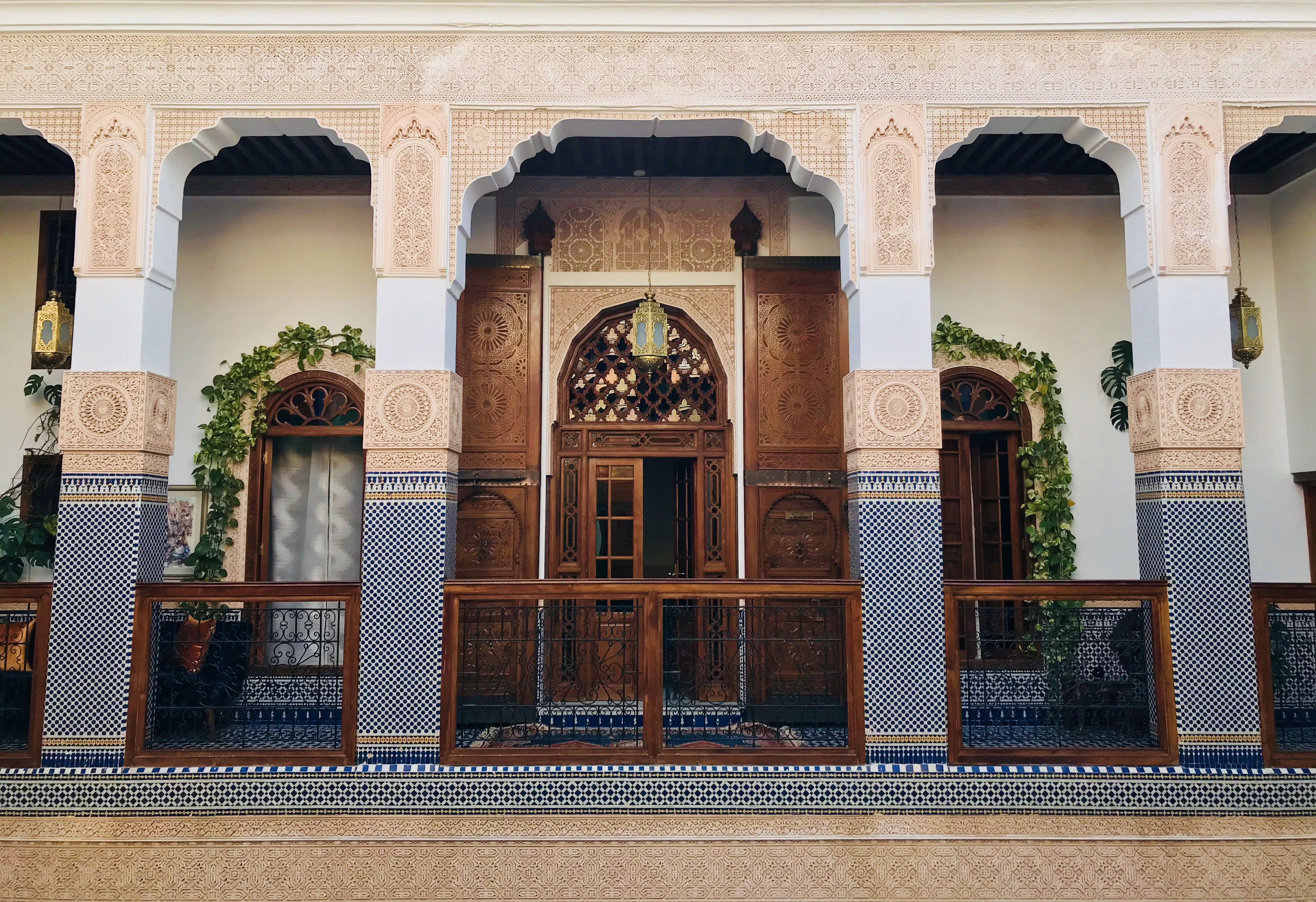 The upper gallery of a riad in the Fez medina: carved plaster arches on white columns, blue-and-white zellij panels, tall cedar doors and brass lanterns around the open courtyard.