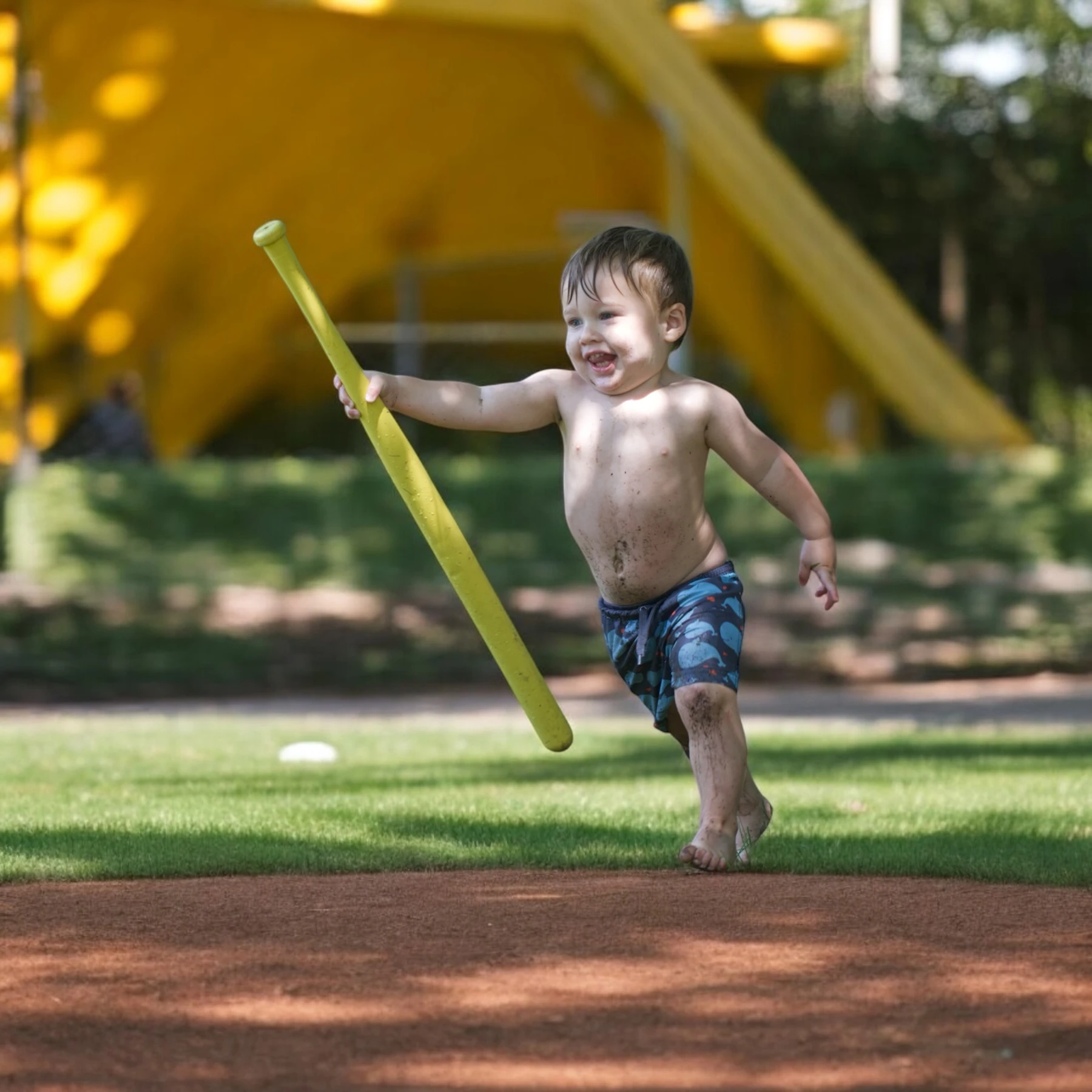 A toddler running across a lawn swinging a long yellow plastic bat, a white ball in the grass