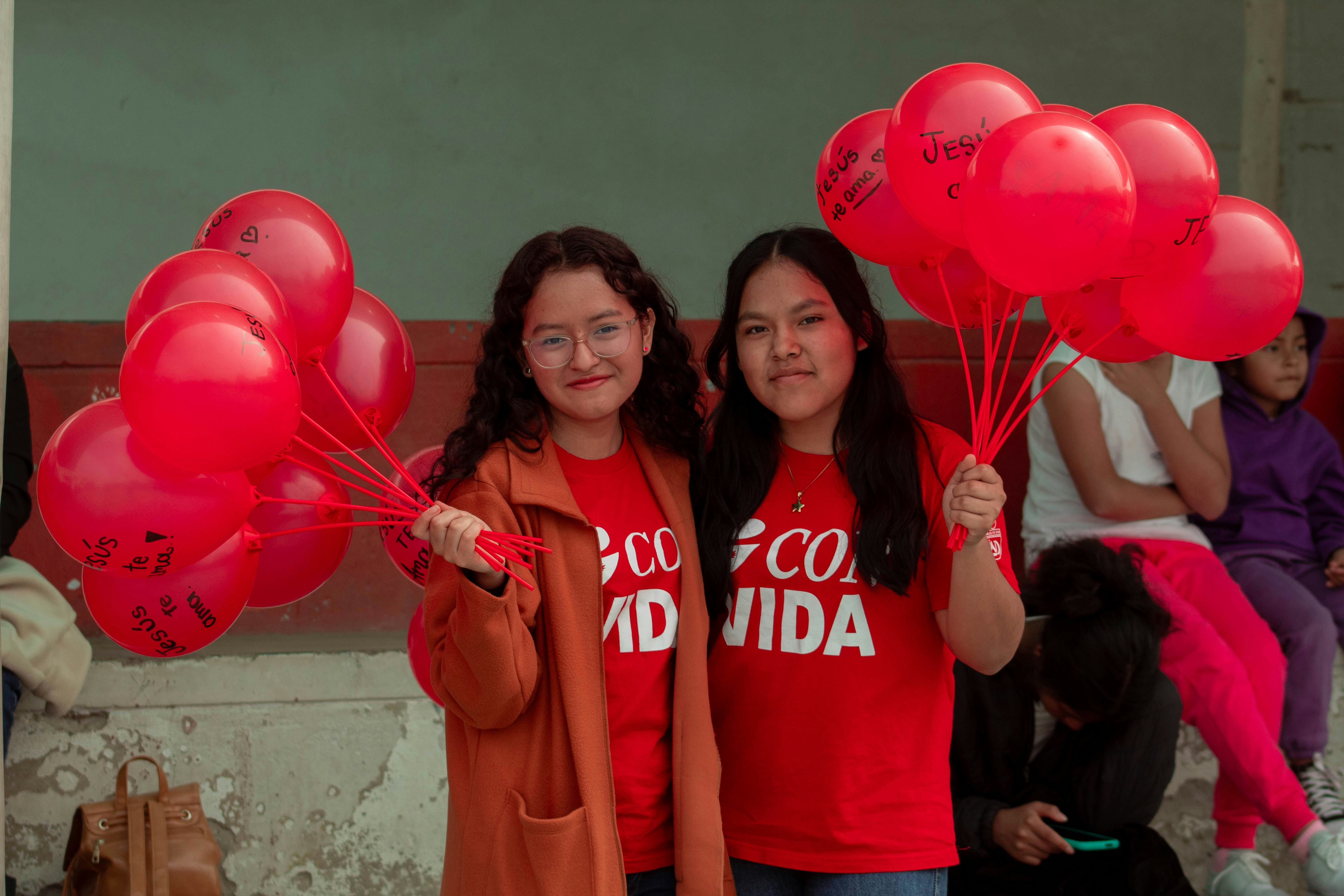 Two girls are holding red heart shaped balloons