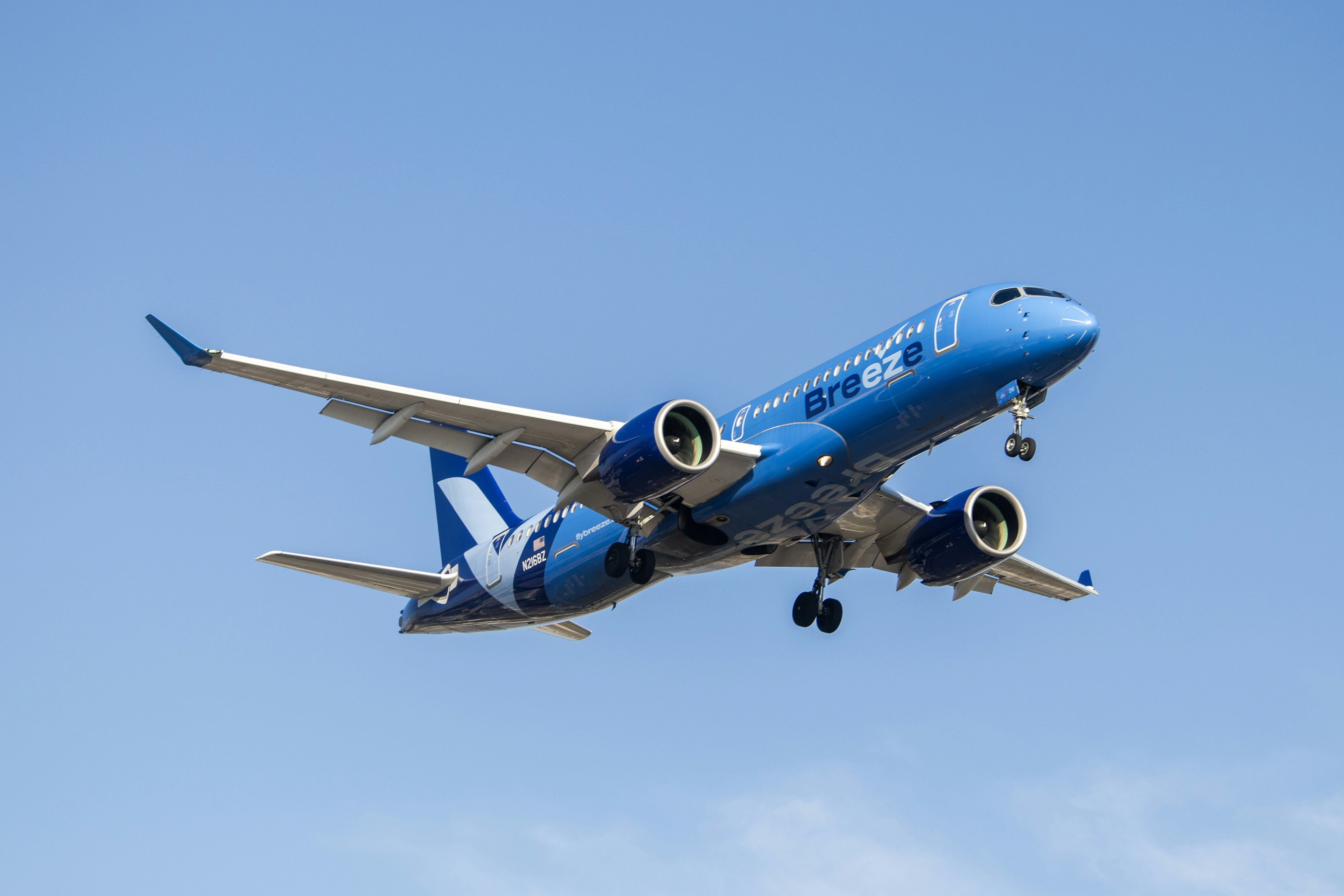 A blue and white jet airliner flying in the sky, An Airbus A220 from Breeze on a landing approach at the John Wayne Airport in Santa Ana, California.