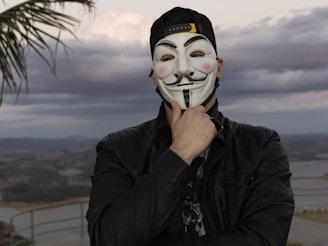 A man wearing a guy fappe mask with a palm tree in the background