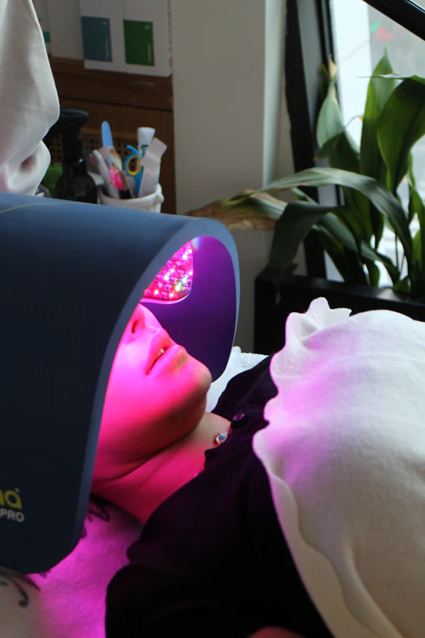 Client reclining beneath a domed light-therapy lamp casting pink light across her face