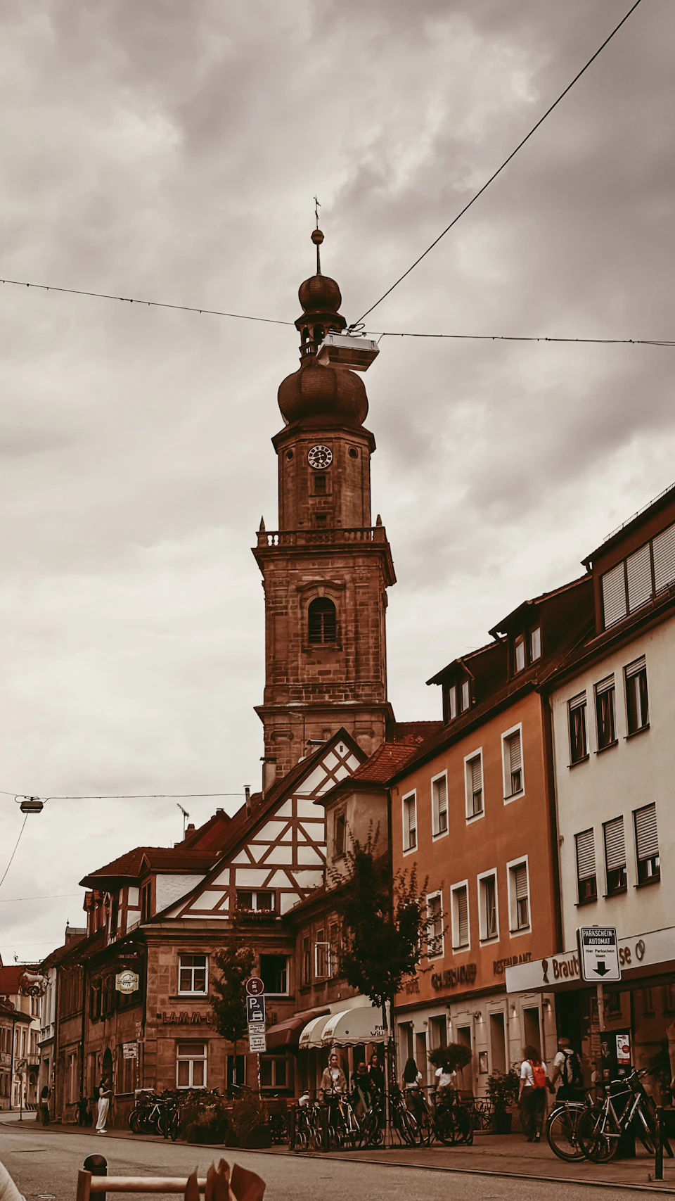 Quaint European town scene features tall brick clock tower against overcast sky prominently