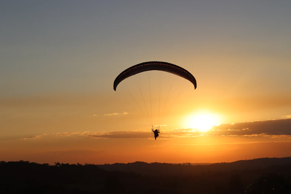 The image depicts a person paragliding over a breathtaking sunset. The paraglider is suspended in mid-air, with their body angled towards the right side of the frame and their feet pointed upwards. Below them, the sky is painted in warm hues of orange and yellow, creating a stunning backdrop for this airborne adventure.

The paraglider's parachute, a black silhouette against the vibrant sunset, is attached to a rope that extends outwards from the center of the image towards the bottom right corner. The scene is set against a serene landscape with rolling hills in the background and trees dotting the horizon line.