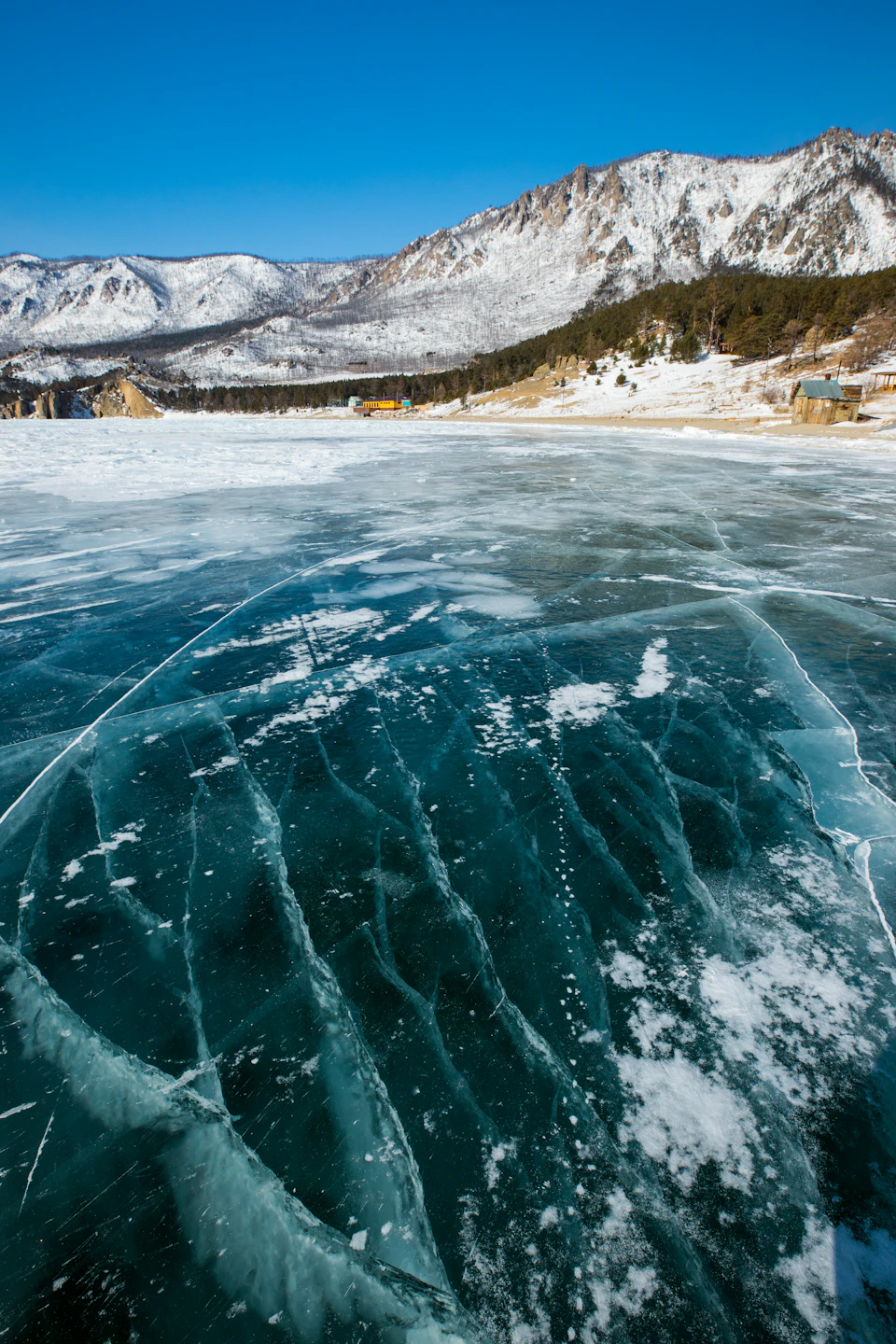 Serene winter landscape with frozen lake, snow-covered mountains, and trees in clear blue sky