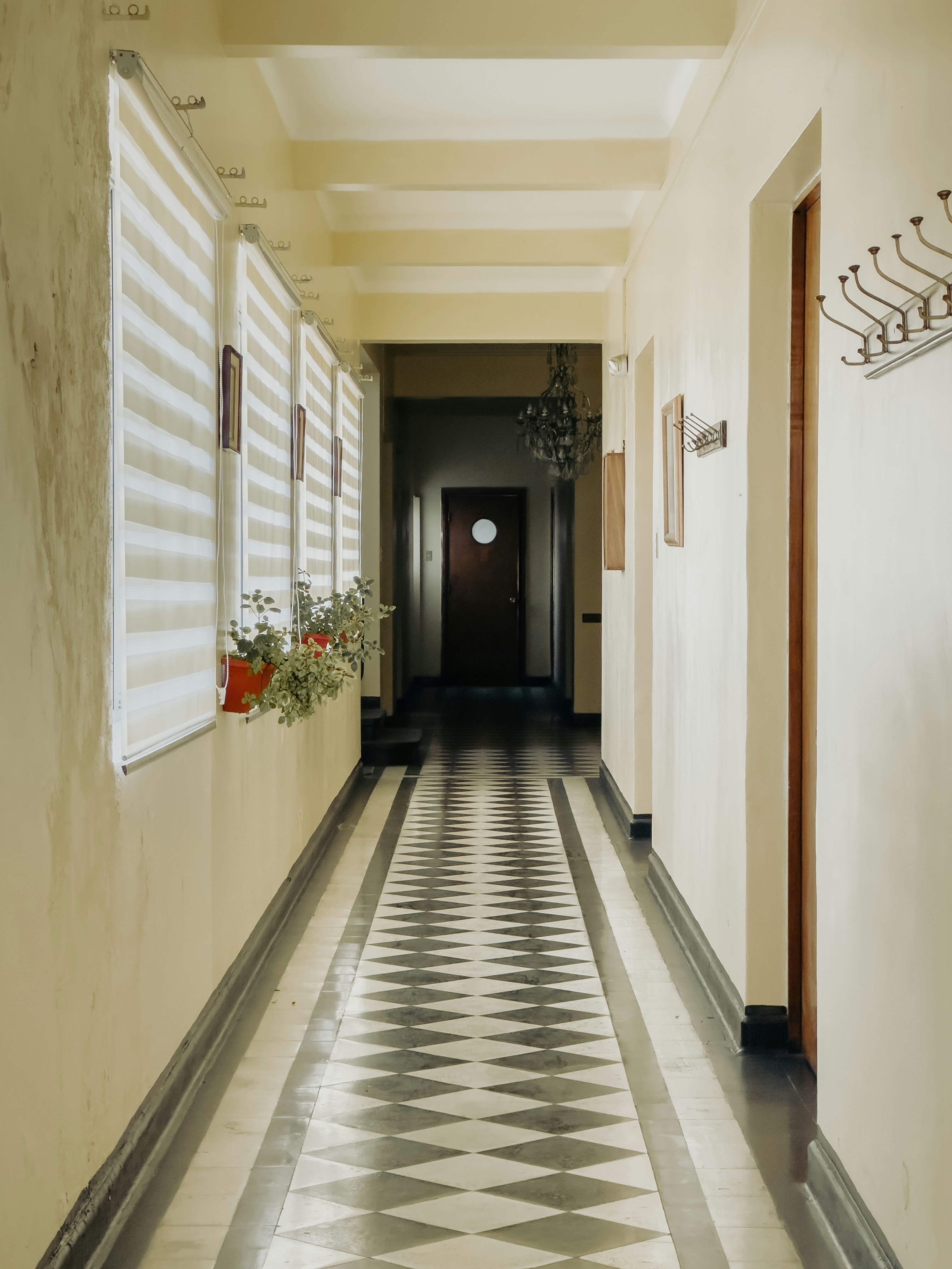 Un pasillo iluminado del Museo Marítimo Nacional de la Armada de Chile en Valparaíso. La imagen muestra un elegante piso de baldosas en blanco y negro con un diseño geométrico, rodeado por paredes de tonos cálidos y ventanas con persianas de luz suave que crean un ambiente tranquilo y nostálgico. Una planta en maceta añade un toque de vida al espacio, mientras que al fondo se observan detalles de una puerta y una lámpara de araña, evocando la rica historia marítima y patrimonial del lugar. Ideal para quienes aprecian la arquitectura y los espacios históricos.