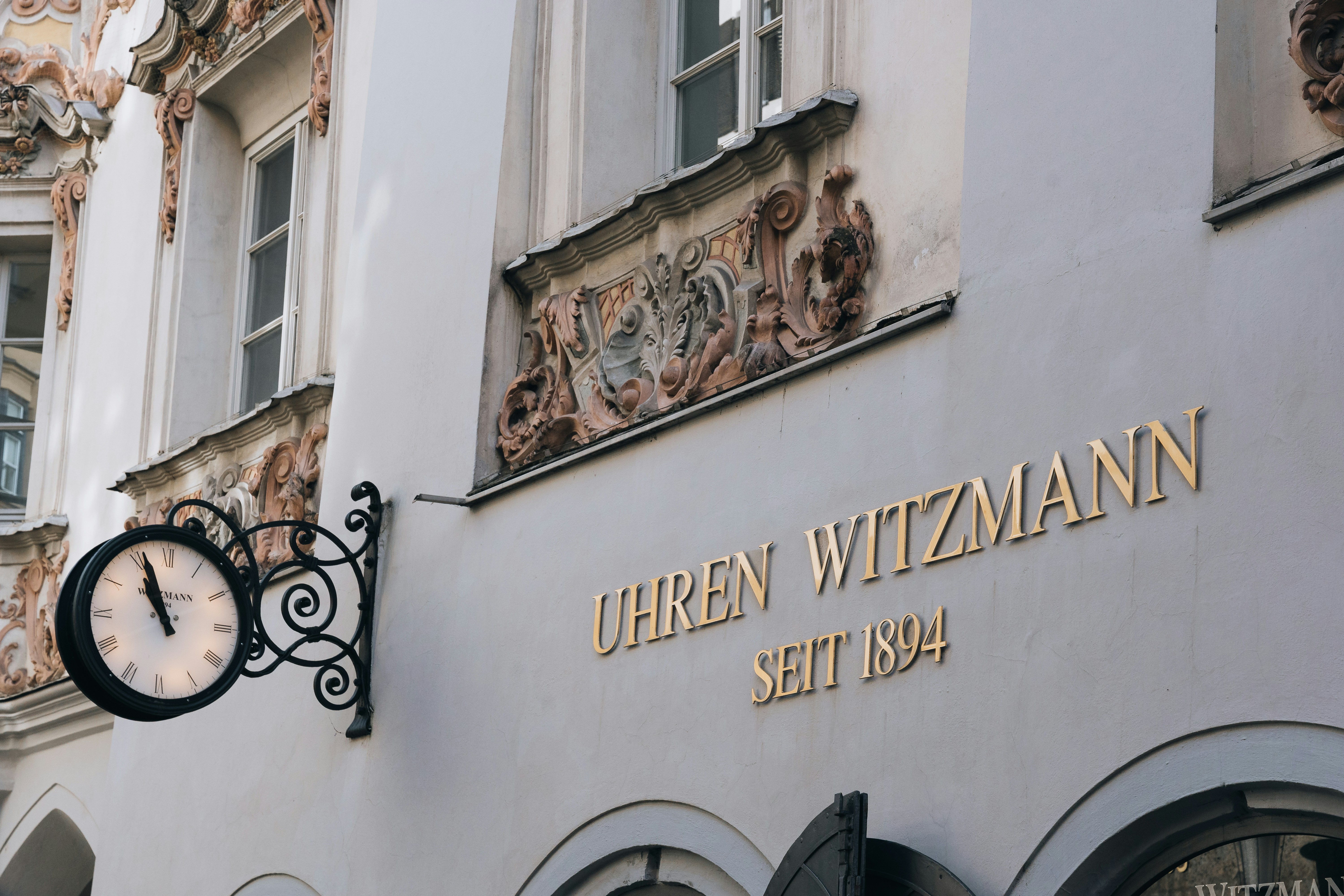 An ornate shop facade features an elegant black clock with Roman numerals, mounted on wrought iron scrollwork beside gold lettering that reads 'Uhren Witzmann, Seit 1894.' Delicate architectural details and faded stucco evoke old-world charm and timeless craftsmanship.