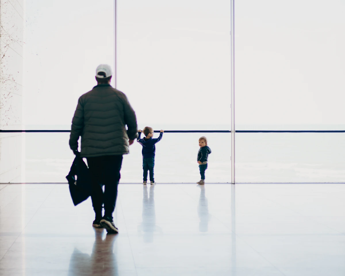 A man carrying a bag walks towards two small children looking out of a large airport window