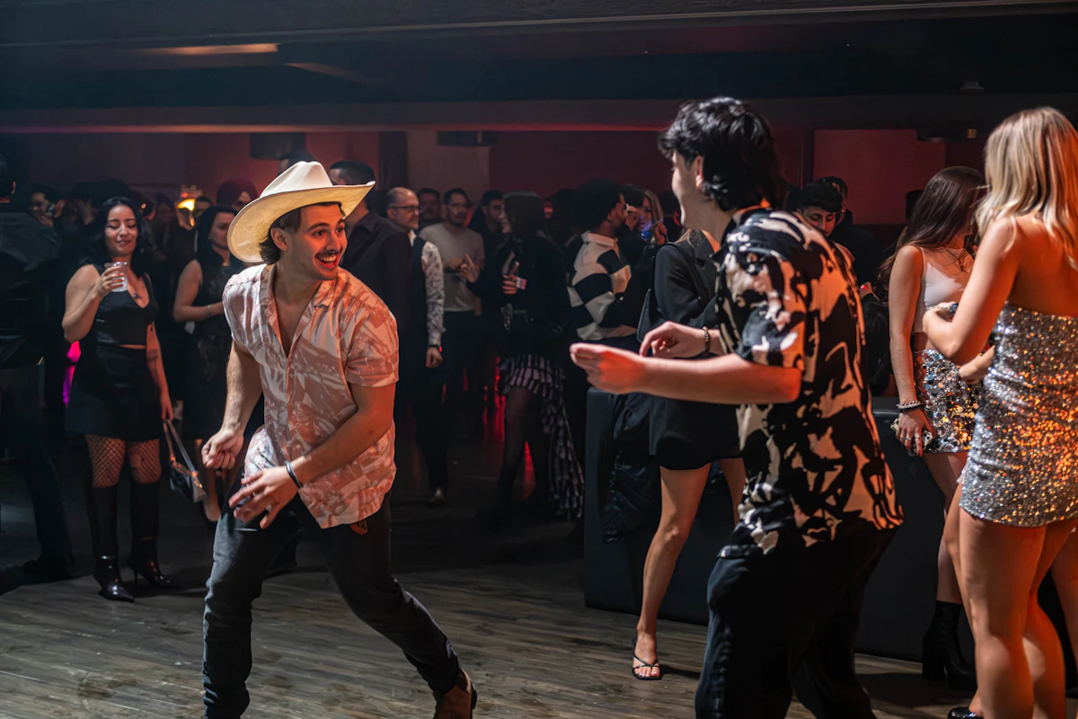 Young adult crowd dancing at a country-western themed venue, one dancer wearing a white cowboy hat with stylish modern fashion