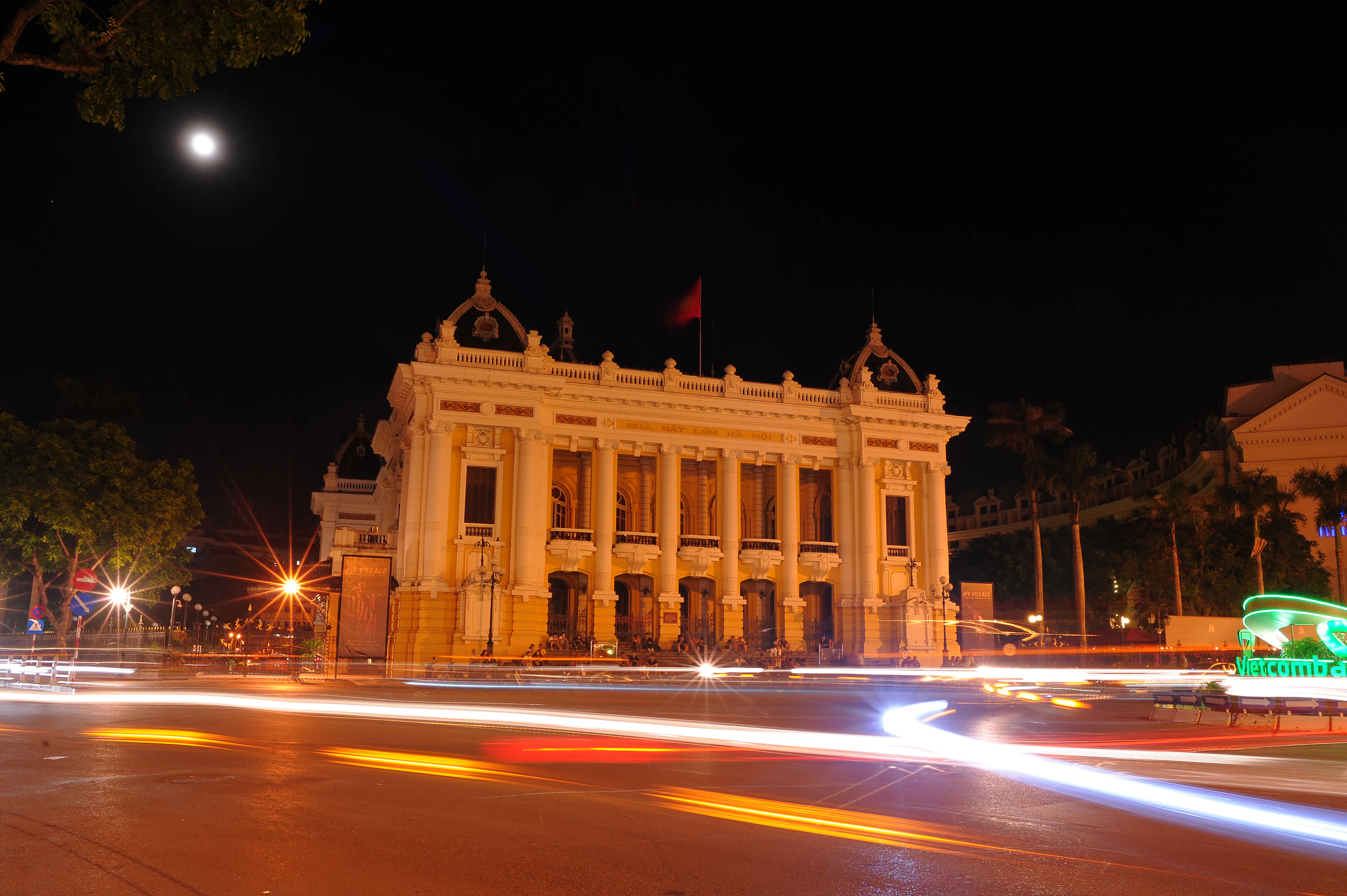 Hanoi's colonial-era Opera House floodlit gold at night beneath a full moon, its columned facade facing a square streaked with the light trails of passing traffic.