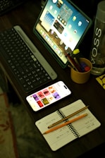 A desk with a laptop, keyboard, phone and a cup of pencils